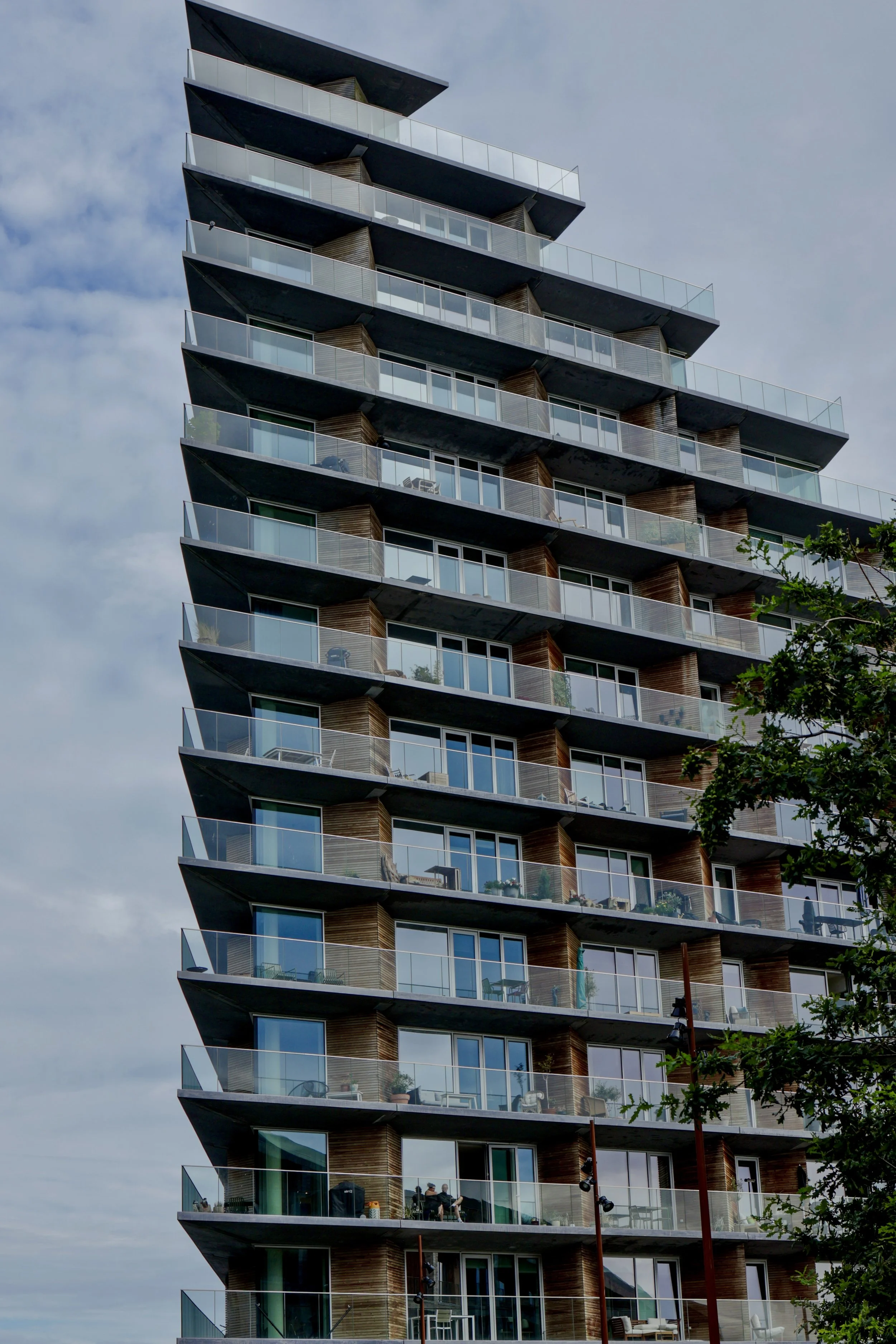 A modern high-rise apartment building with glass balconies and wooden panel accents, set against a partly cloudy sky.
