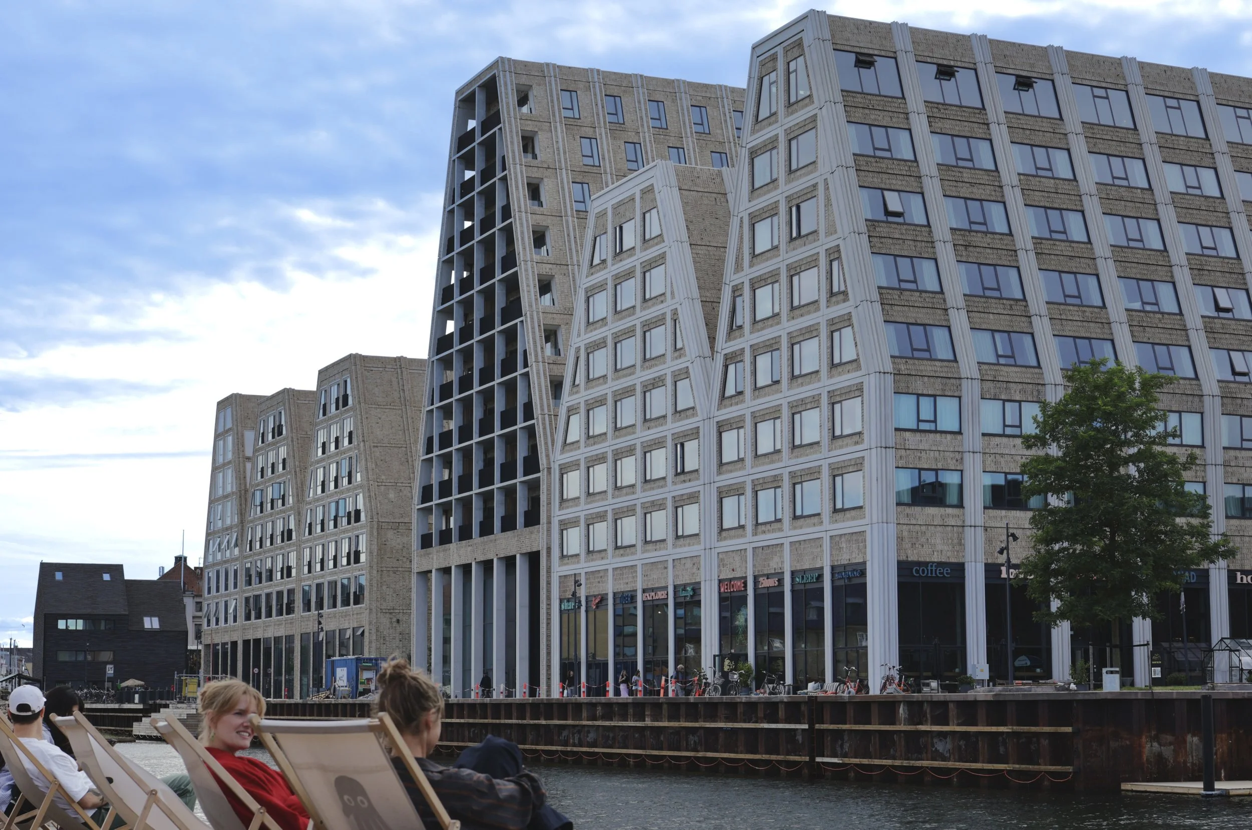 People sitting on deck chairs along a waterway with modern, uniquely shaped apartment buildings in the background, and a cloudy sky above.