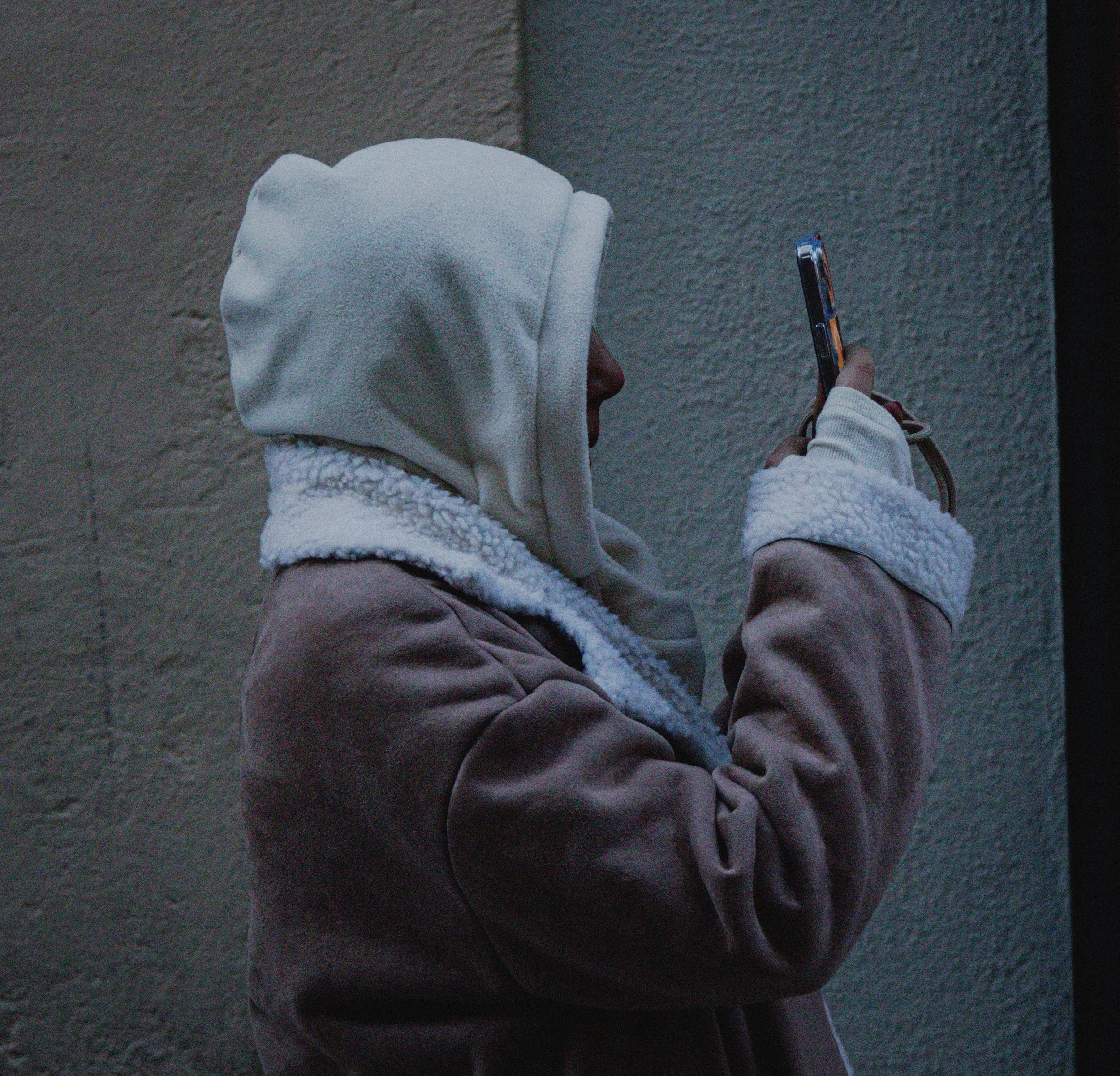 A person wearing a hoodie and brown jacket looking at a smartphone against a gray wall.