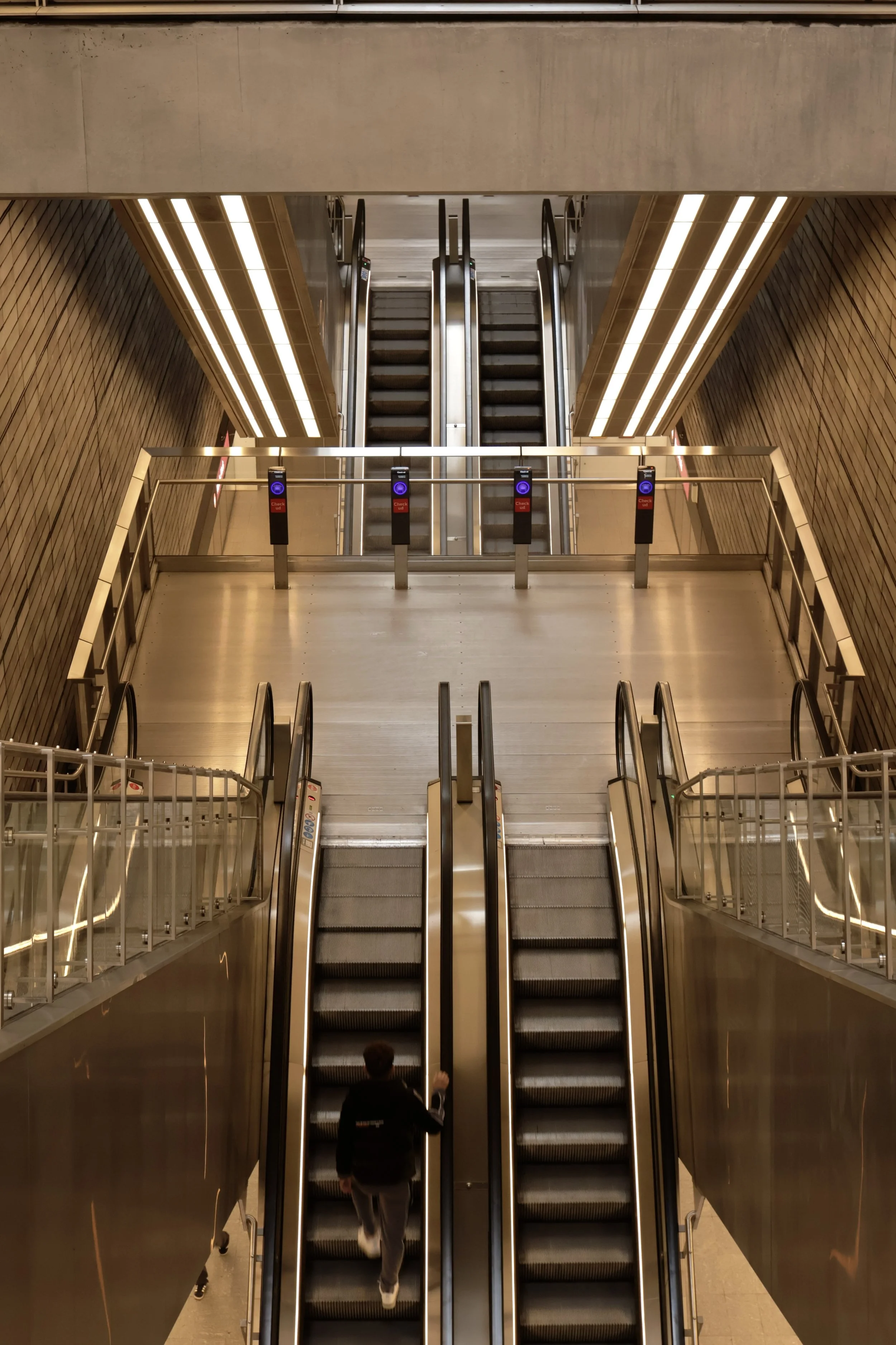 A person in a black hoodie and gray pants ascending escalators in a modern, spacious subway or train station with metallic and brick interior design.