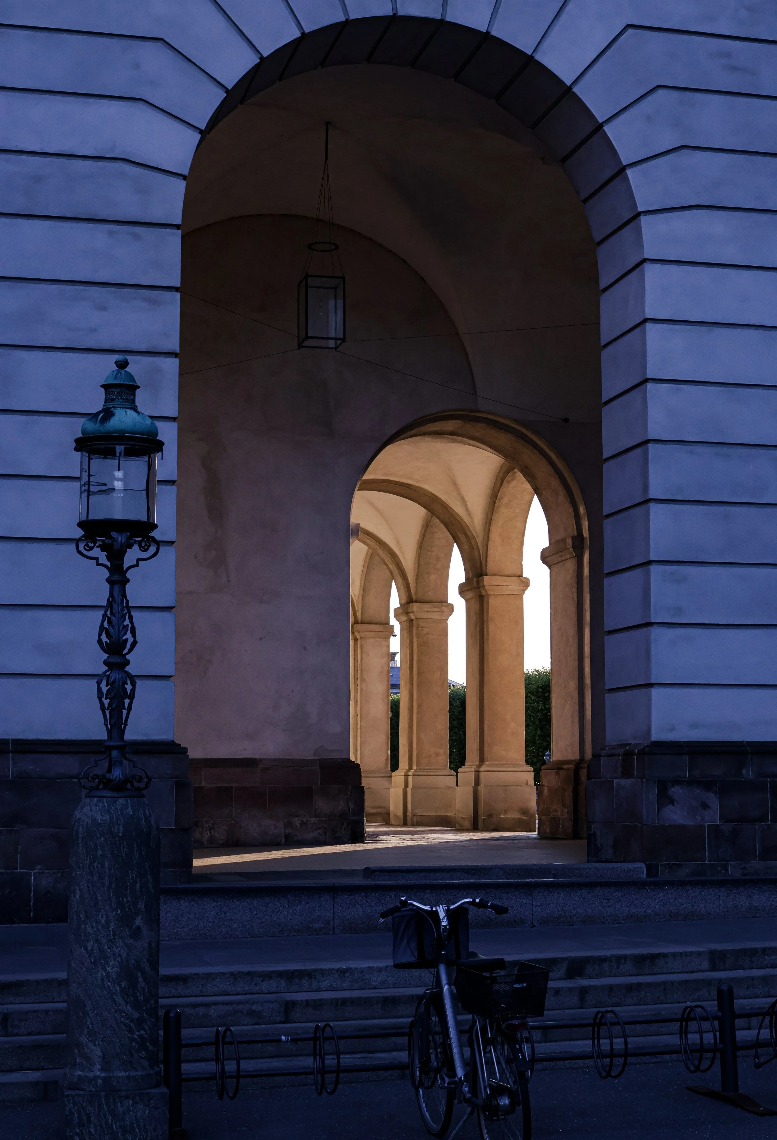 Arched passageway with stone columns, a bicycle parked in the foreground, and a lamppost on a stone pedestal.