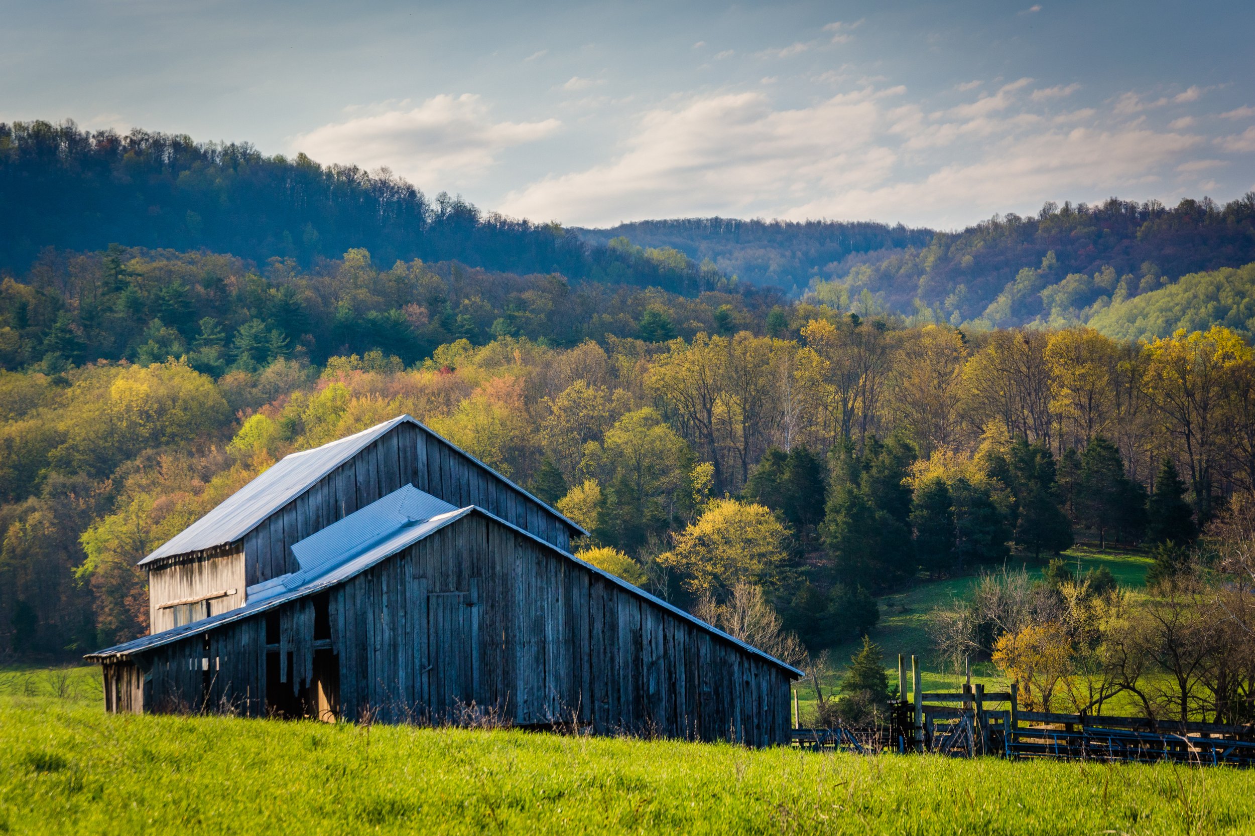 An old wooden barn with a metal roof in a green valley surrounded by hills and trees.