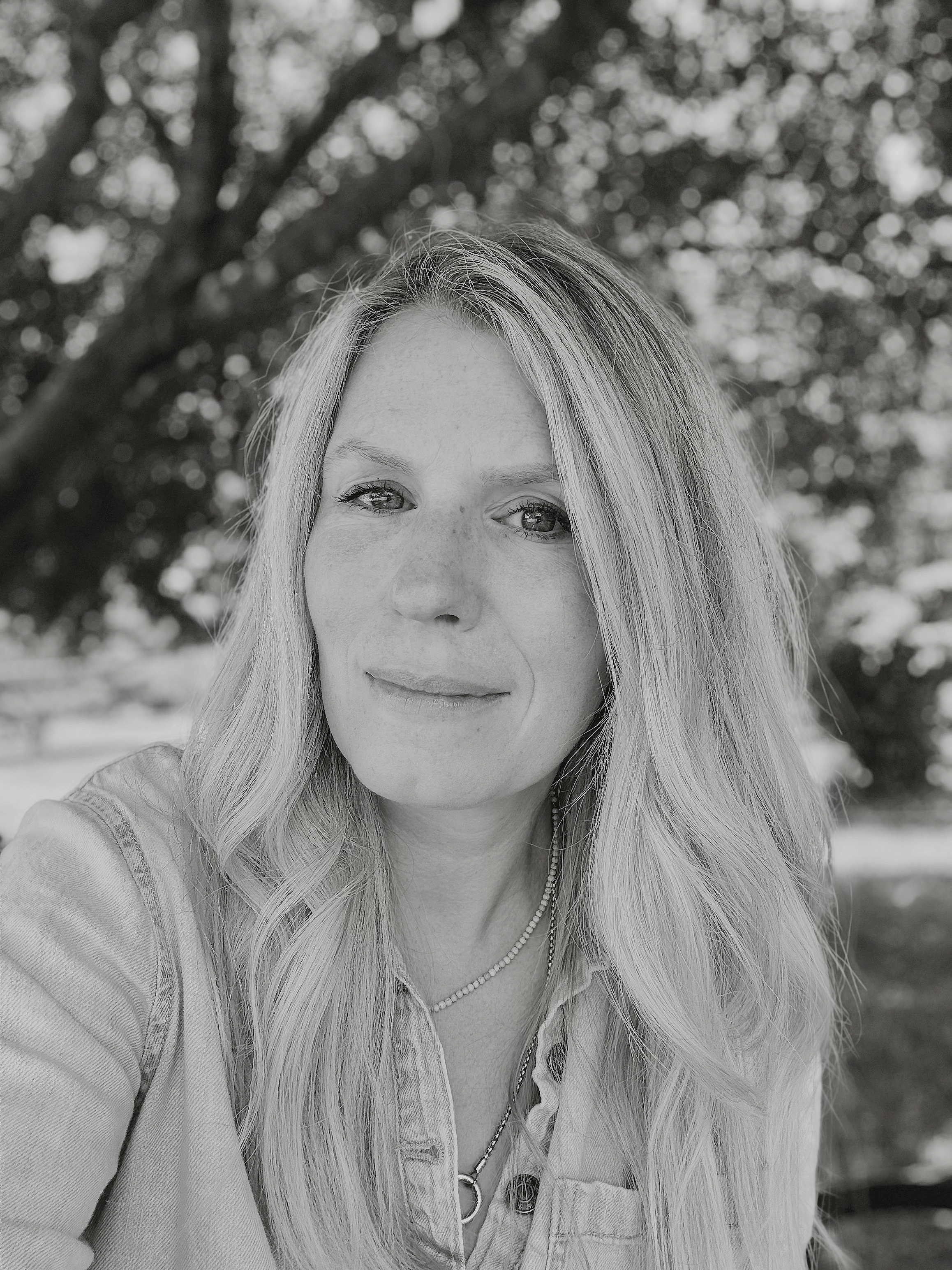 A black-and-white photo of a woman with long wavy hair outdoors in front of a tree.