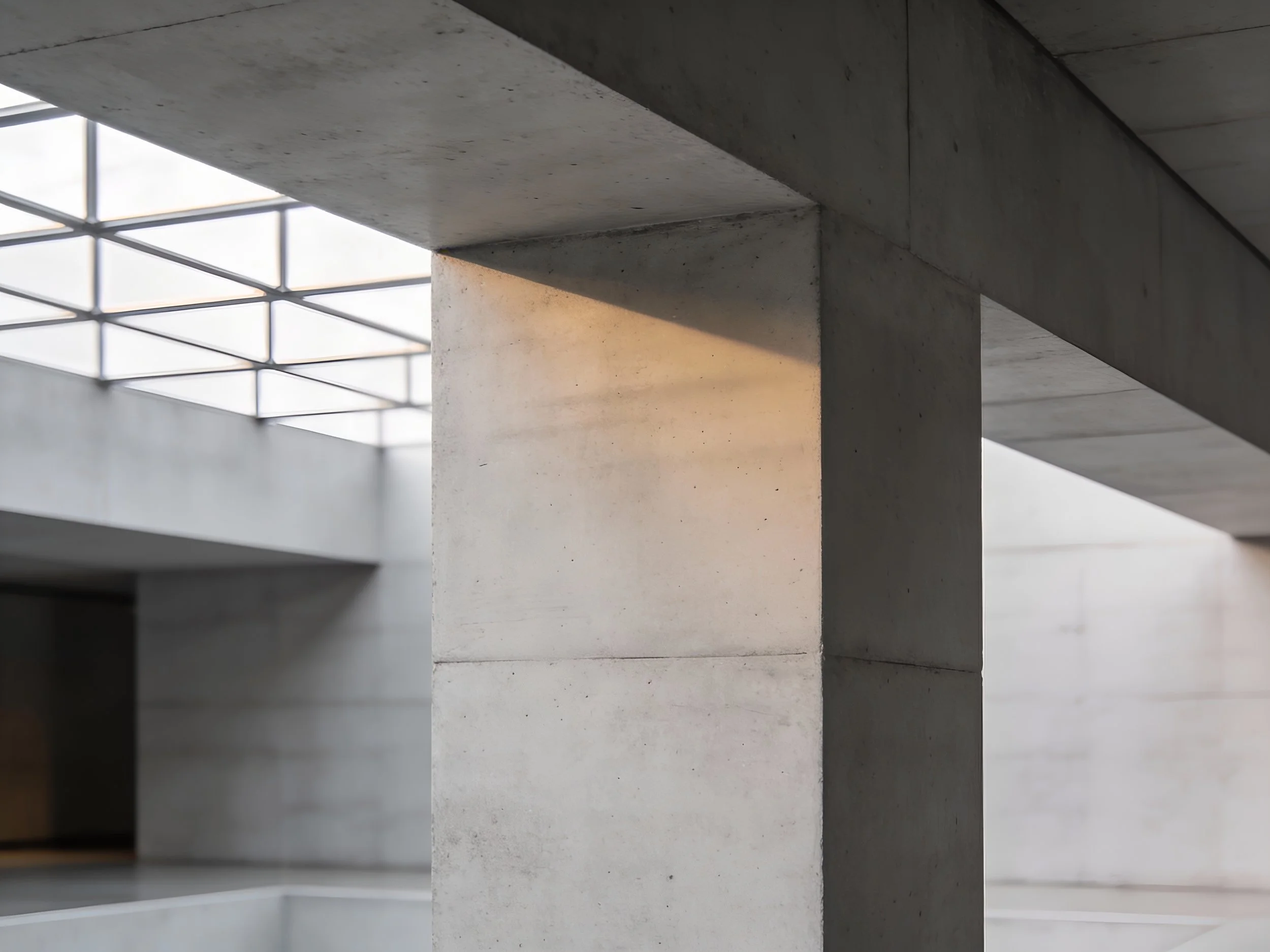 View of a modern building interior with concrete pillars and a grid-patterned skylight, sunlight casting a glow on the pillar.