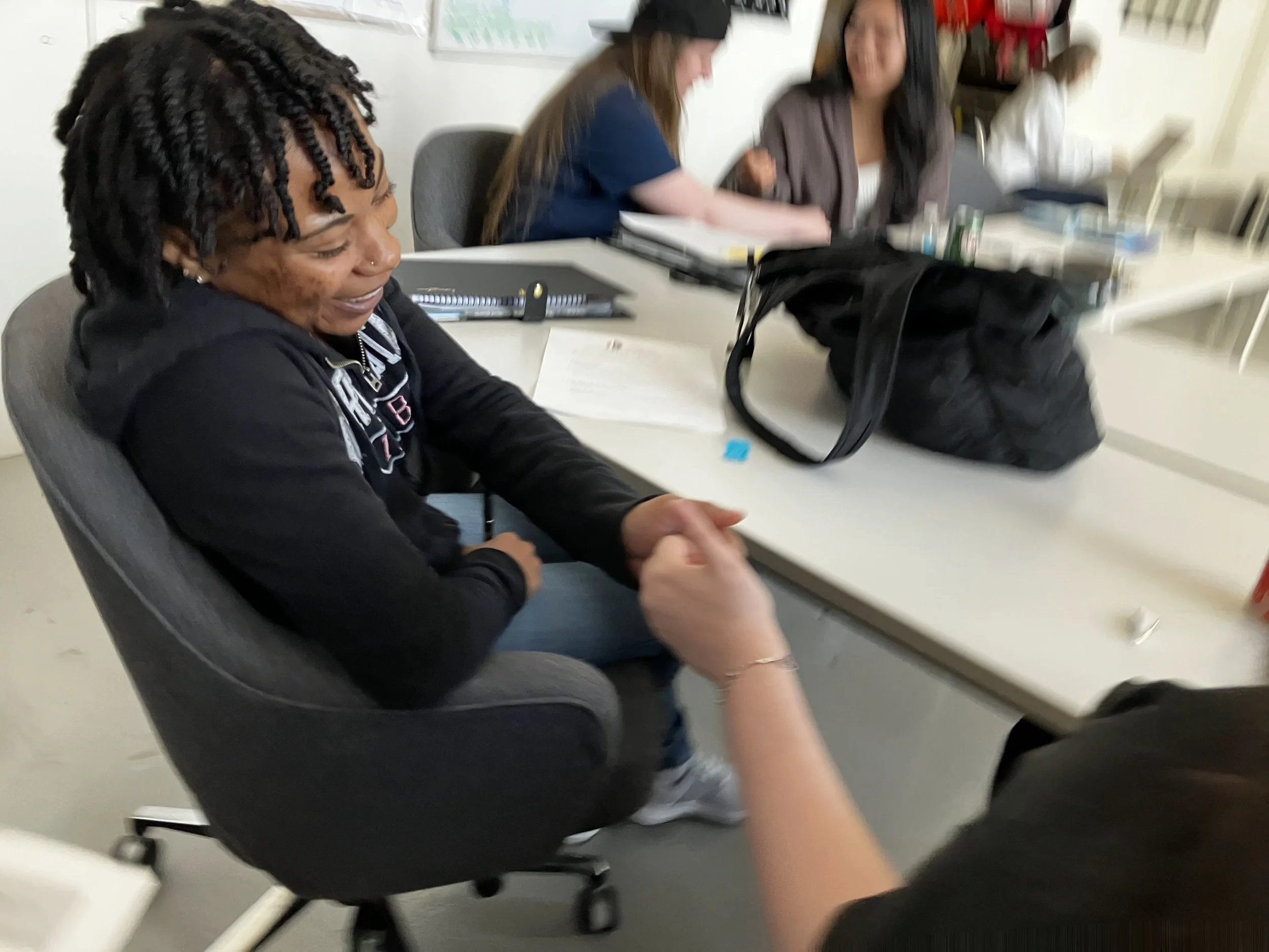 A woman with short, curly hair and a nose piercing is smiling while sitting in a gray office chair at a white table. She is wearing a black hoodie with white and red text, and appears to be engaged in a conversation or activity with someone not fully visible. In the background, two women are sitting at the table with various notebooks and items, and there is a black bag on the table.