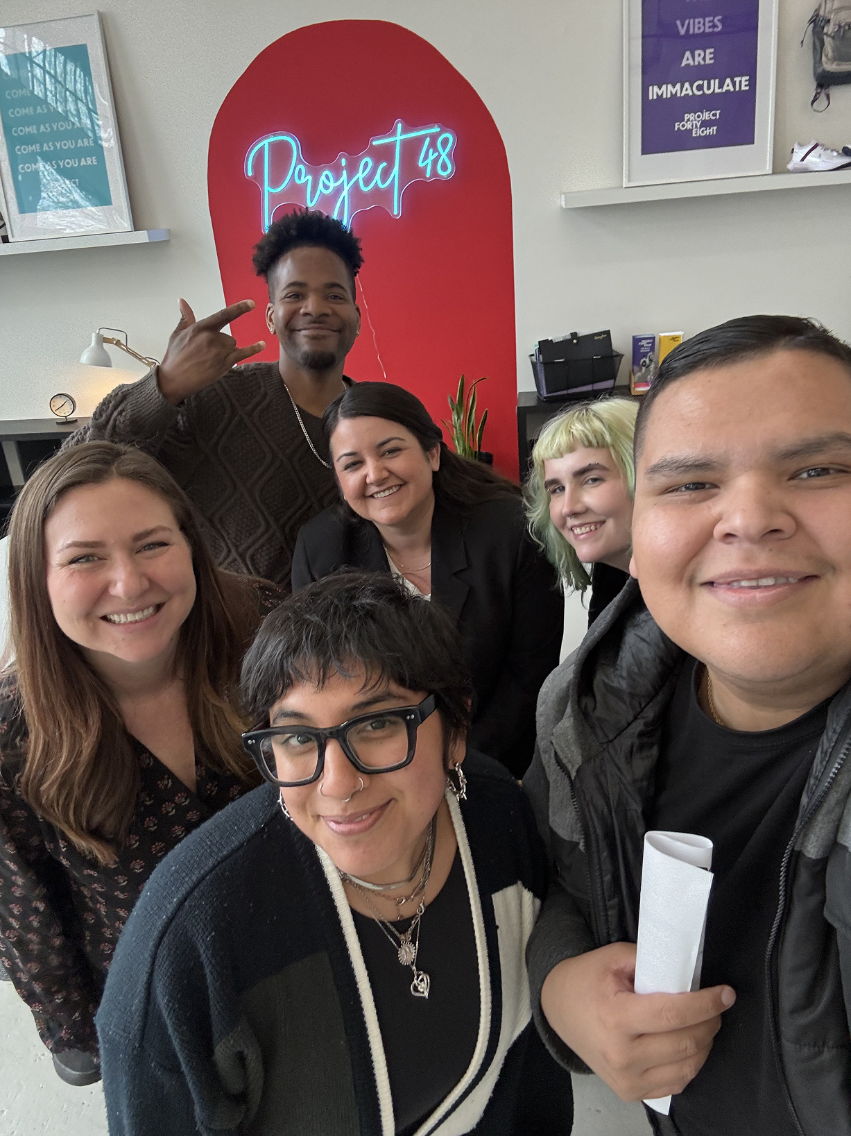 Group of six smiling people taking a selfie in front of a red wall with a neon sign reading "Project 48" during a social event.