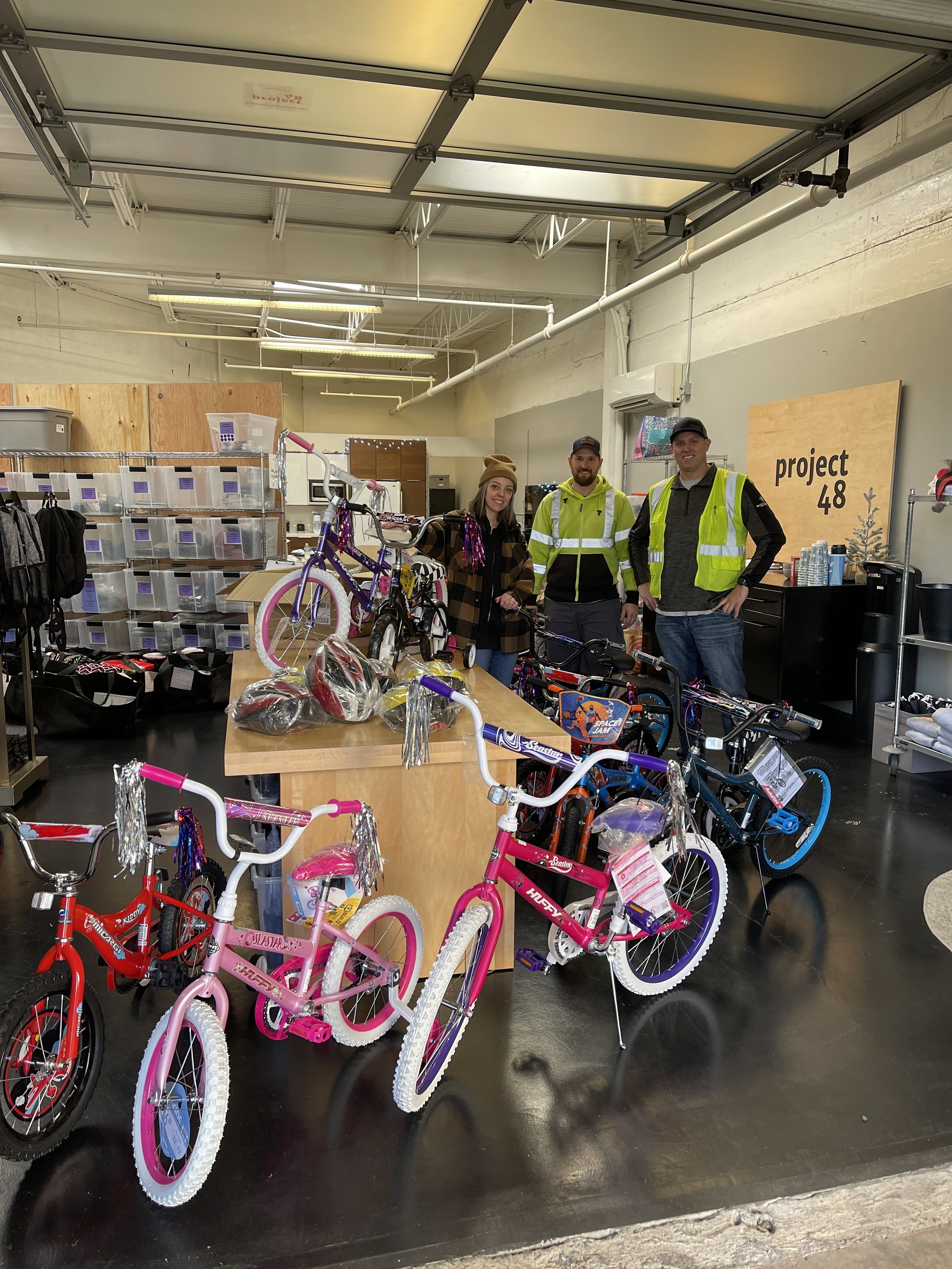 People standing inside a bike shop with several bicycles on display, including pink and red bikes in the foreground, and a table with accessories. There is a sign that reads 'project 48' on the wall.