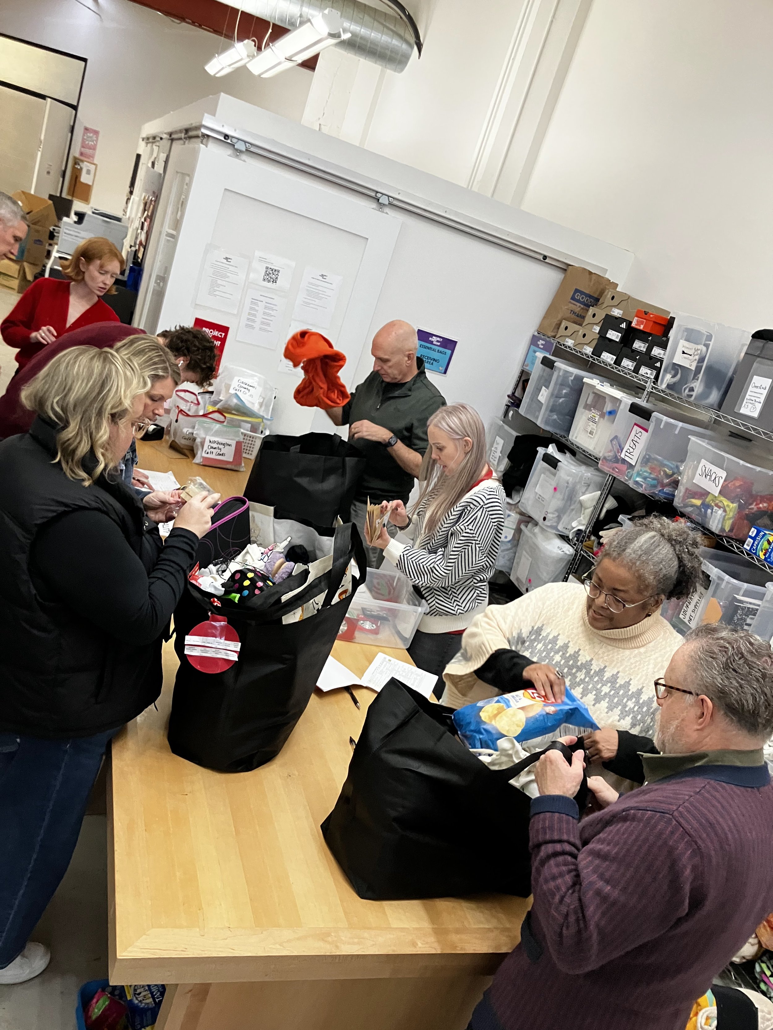 People gathered around a table packing bags, with shelves of supplies in the background, in what appears to be a community or volunteer event.