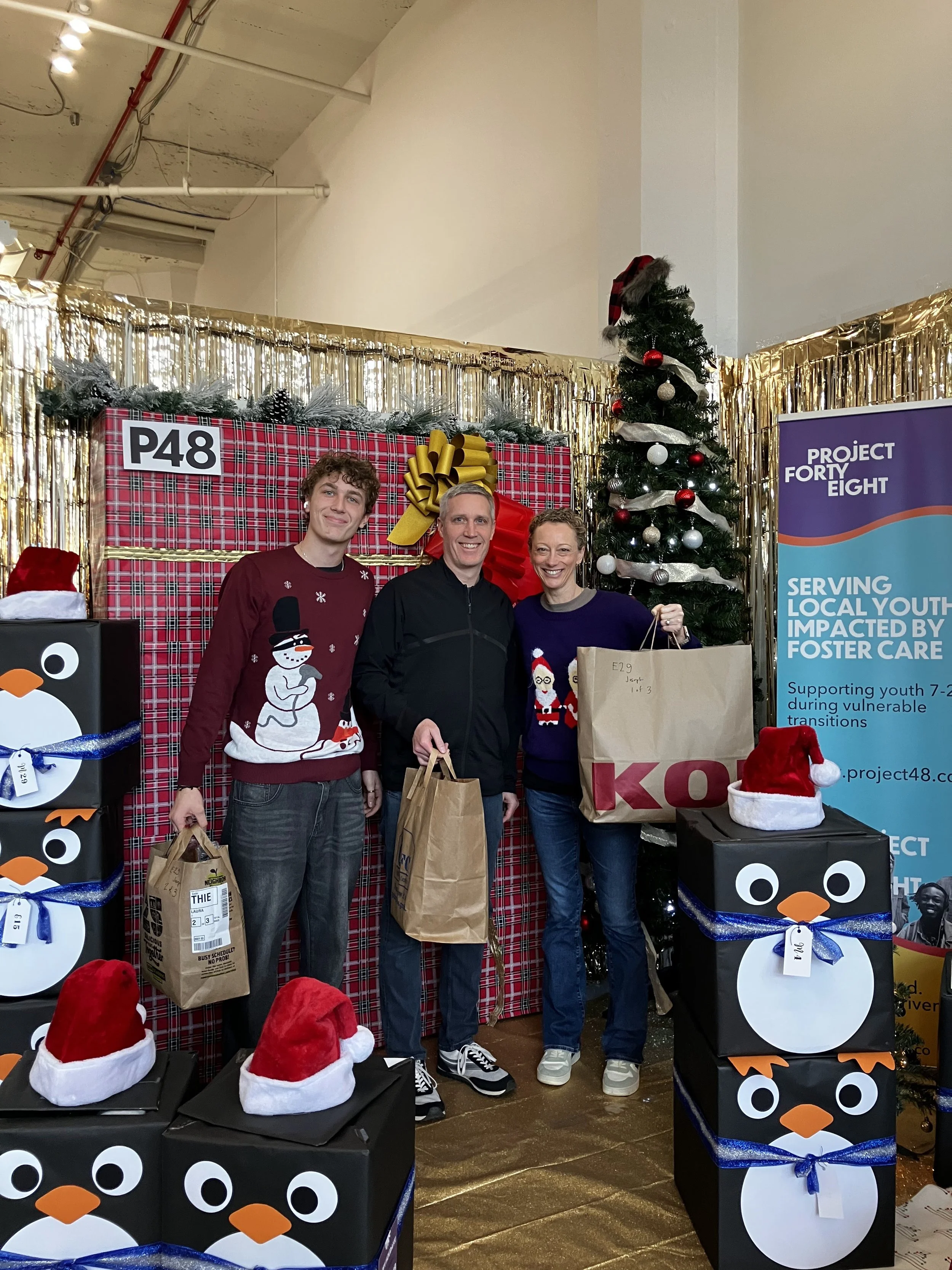 Three people standing in front of a decorated Christmas backdrop with a Christmas tree, gift boxes, and a banner from Project Forty Eight, holding bags of gifts and wearing holiday sweaters.
