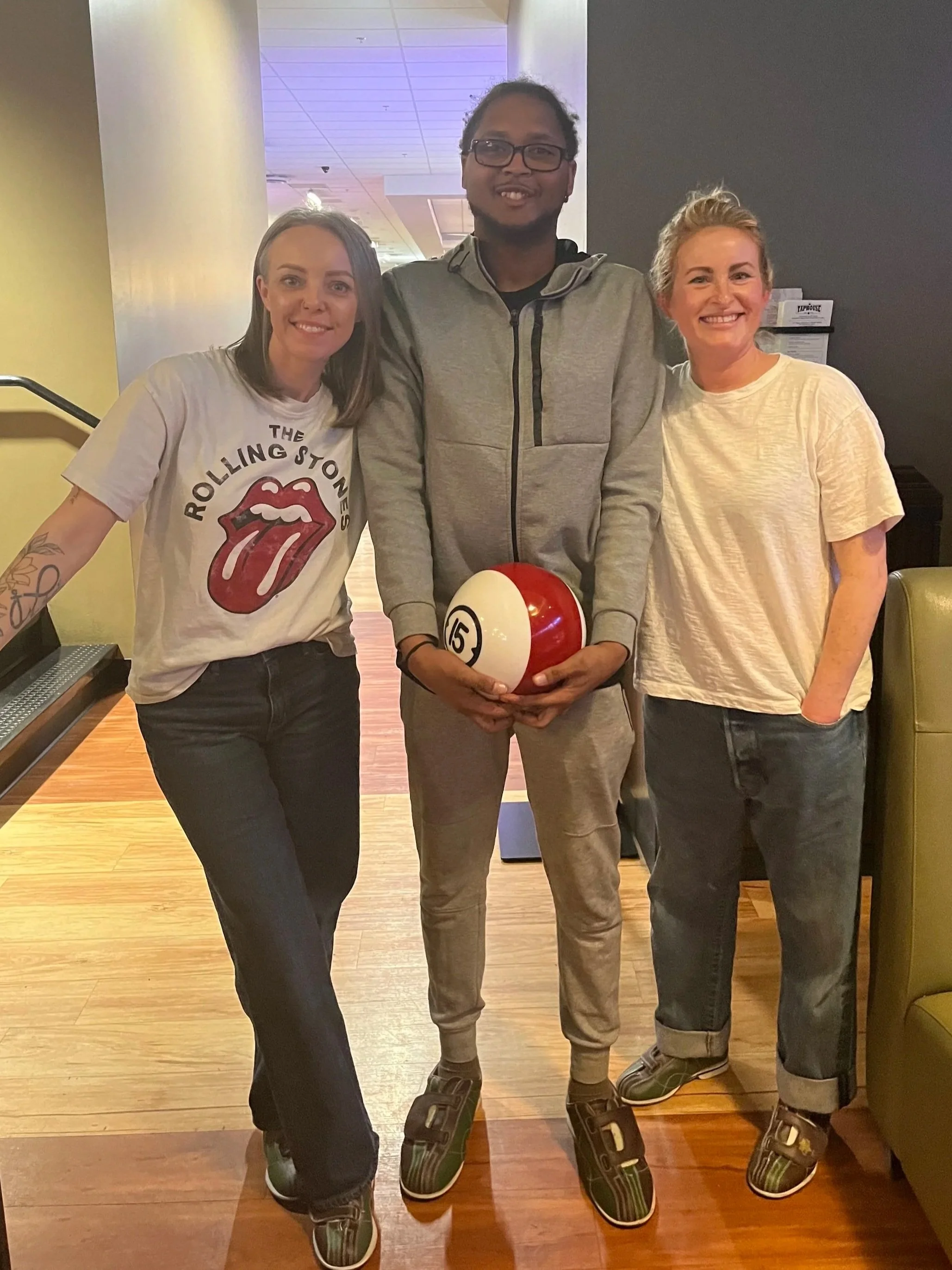 Three women and a man standing together indoors, with the man holding a large red, white, and black 15-ball from a pool table game.