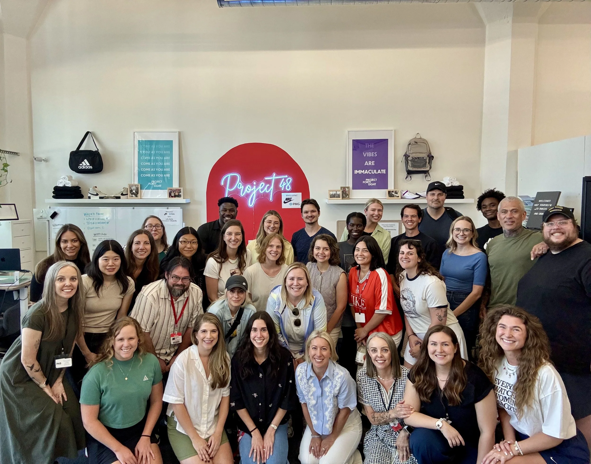 A large group of diverse smiling people gathered in an indoor office space, some standing and some kneeling, in front of a red wall with a neon sign that says 'Project' and a whiteboard with notes. The background features framed posters, shelves with bags, and various decorations.