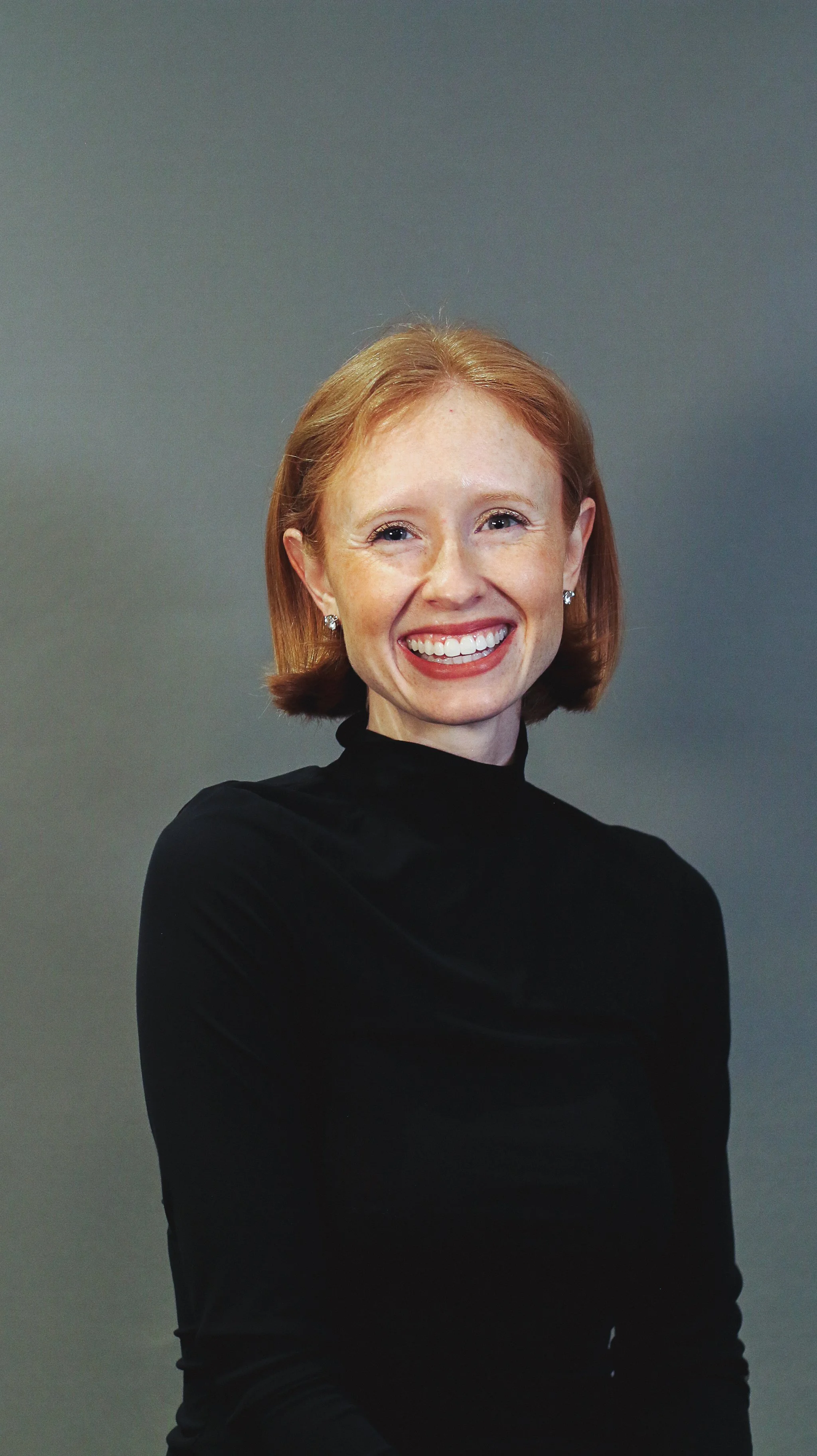 A smiling woman with red hair, wearing a black turtleneck, against a plain gray background.