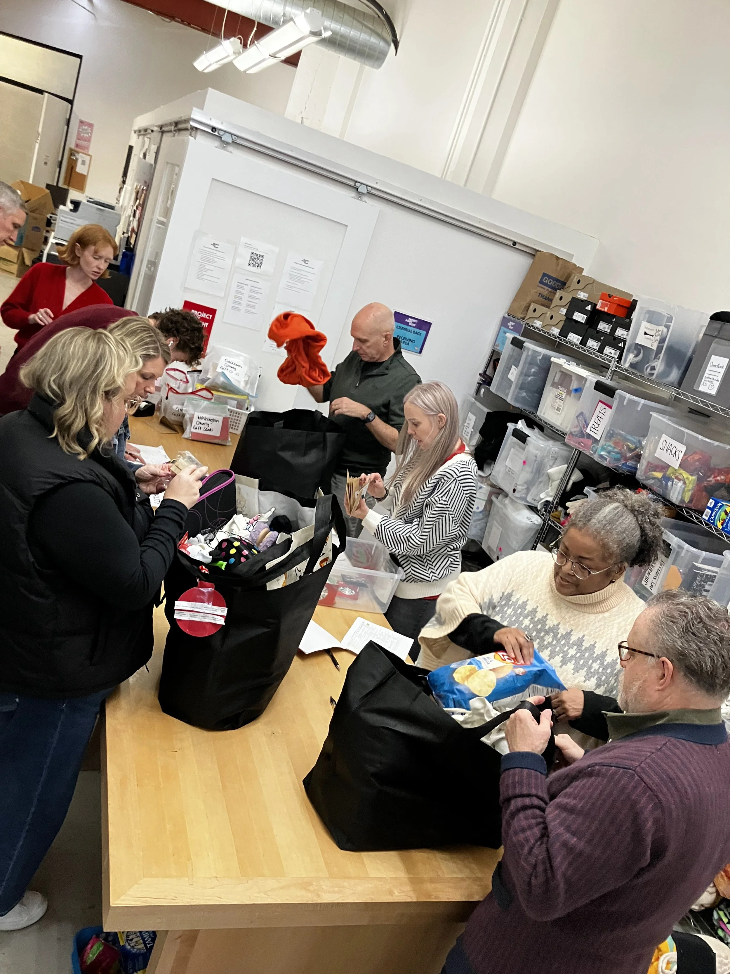 Group of people working together at a table, packing or sorting items in plastic bags, inside a storage room with shelves of labeled containers and boxes.