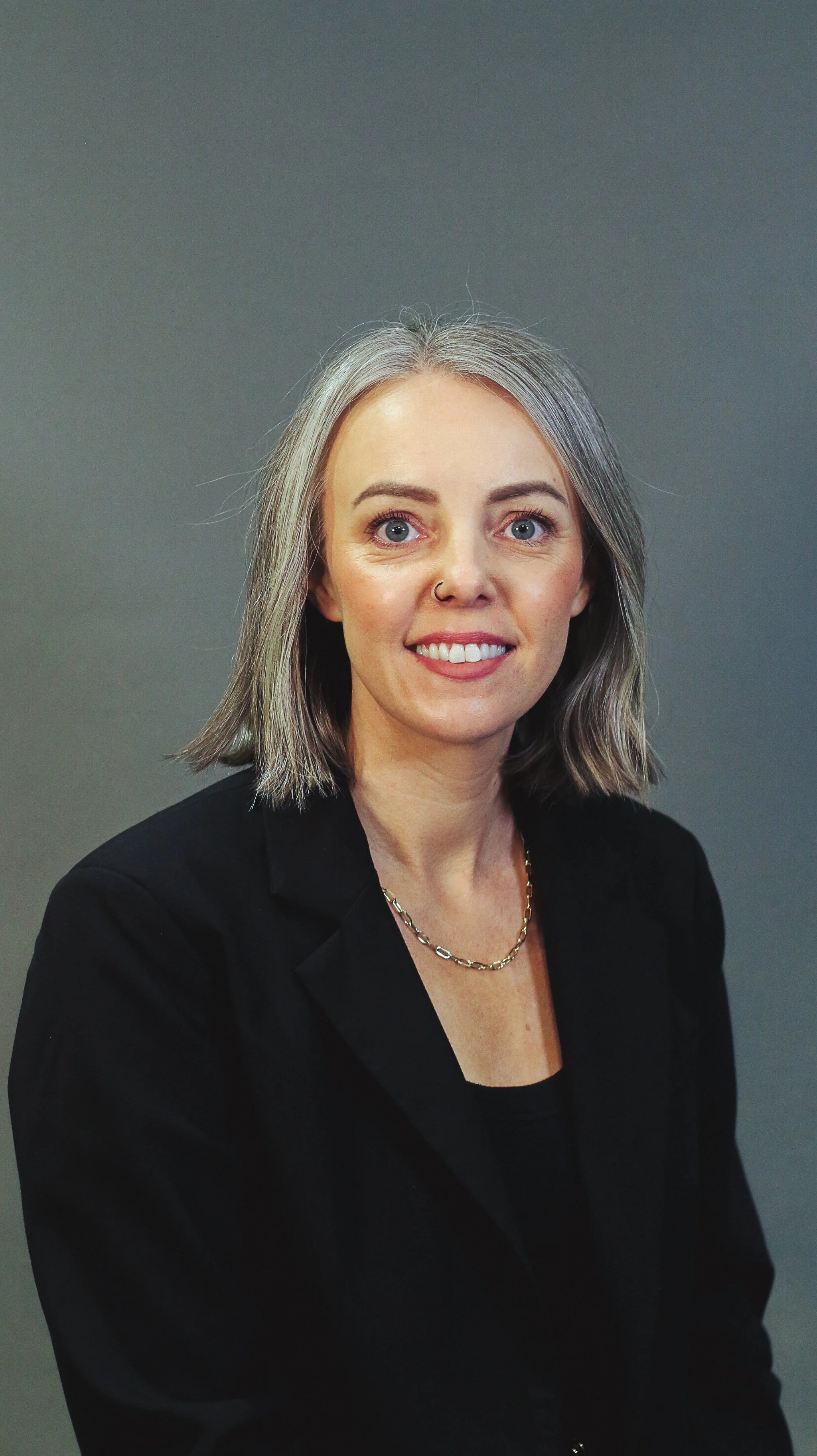 Portrait of a woman with shoulder-length gray hair, blue eyes, wearing a black blazer and a gold chain necklace, smiling against a plain gray background.