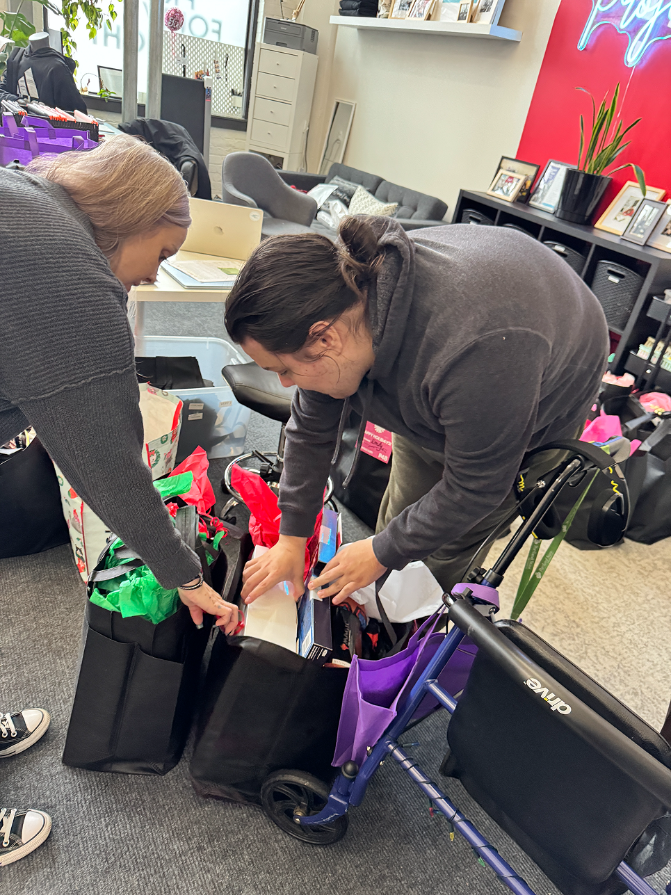 Two women are arranging gift bags and items in black shopping bags inside an office or store space. There are various items, a purple cart, and shelves with photographs and decor in the background.