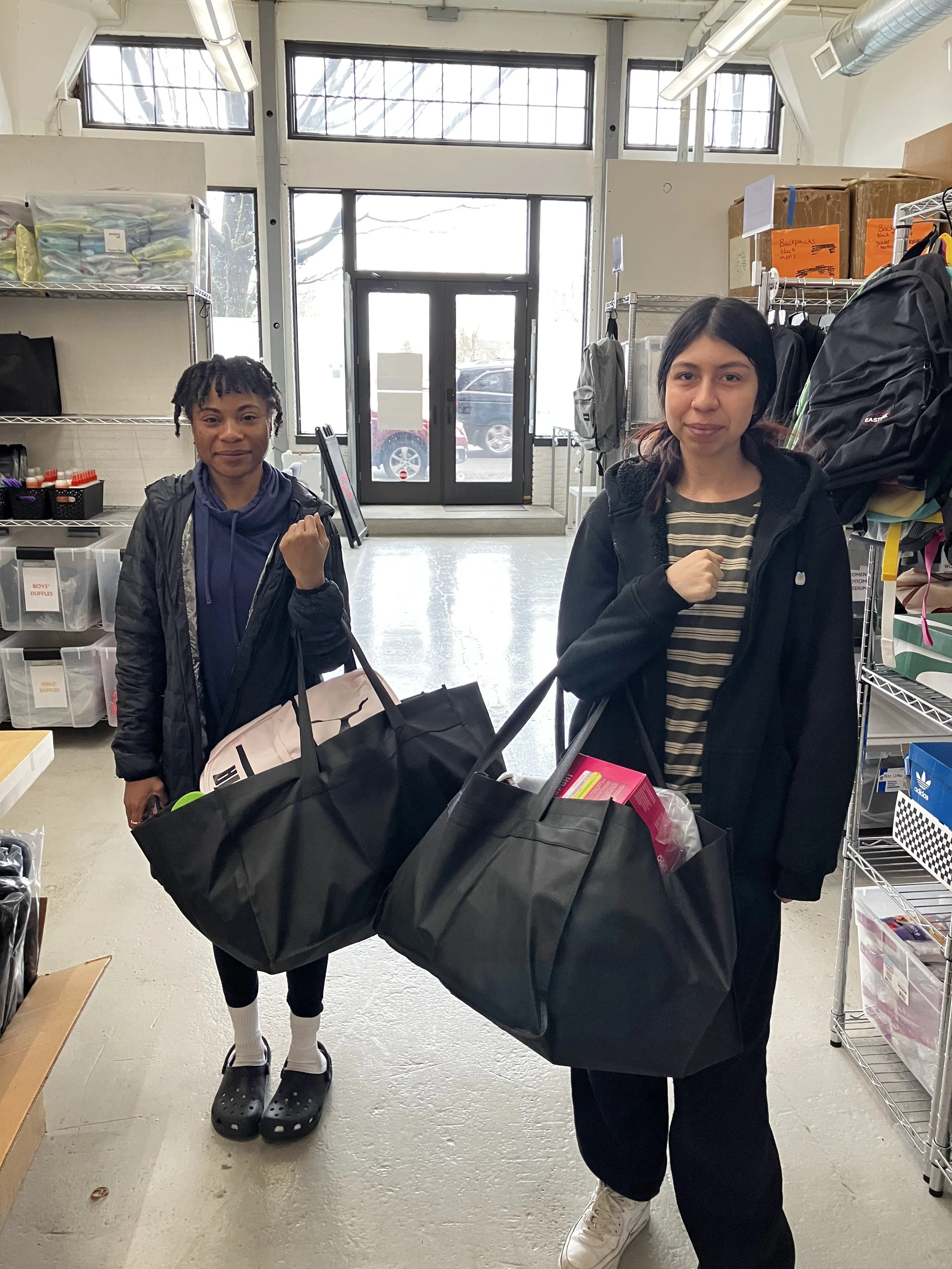 Two women standing inside a store, each holding large black bags filled with items. The woman on the left is wearing a black jacket, blue hoodie, black pants, white socks, and Crocs. The woman on the right is wearing a black jacket, striped shirt, black pants, and white sneakers. Behind them are shelves with various supplies and a glass door at the back of the store.