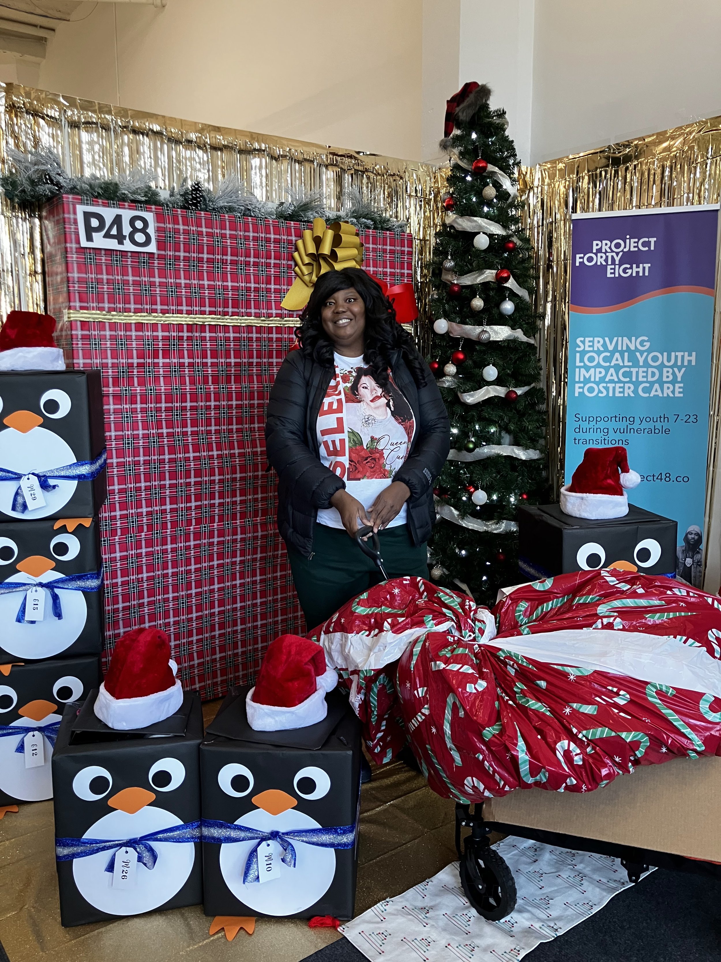 A woman standing in front of a decorated Christmas display with Christmas presents, a decorated Christmas tree, and a woman smiling, holding a gift wrapped in red and green holiday wrapping paper, with penguin-themed boxes and Santa hats around her.