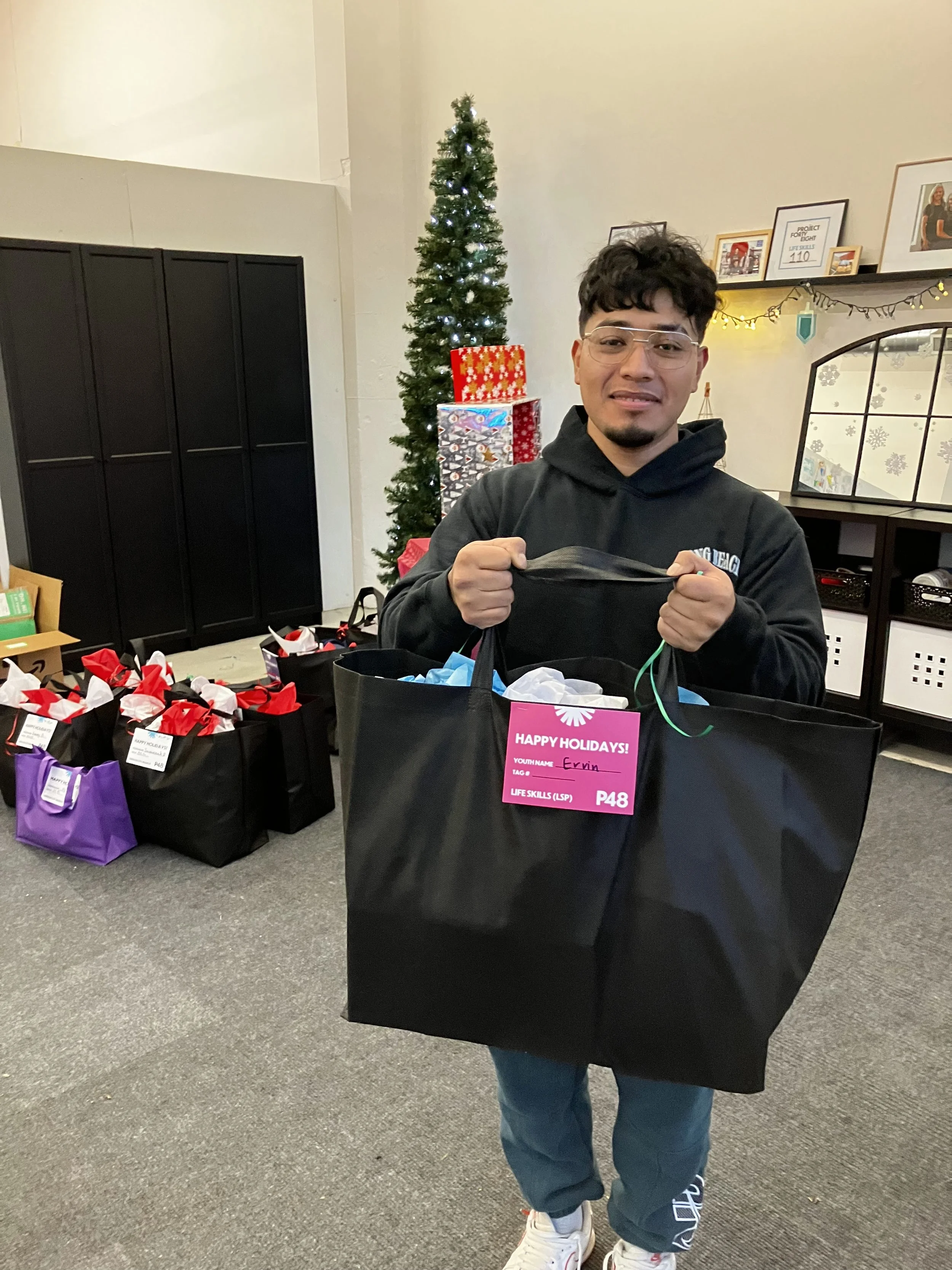 Young man holding a large black gift bag with a pink label that reads 'Happy Holidays', standing near a decorated Christmas tree and other gift bags in a room decorated for the holidays.