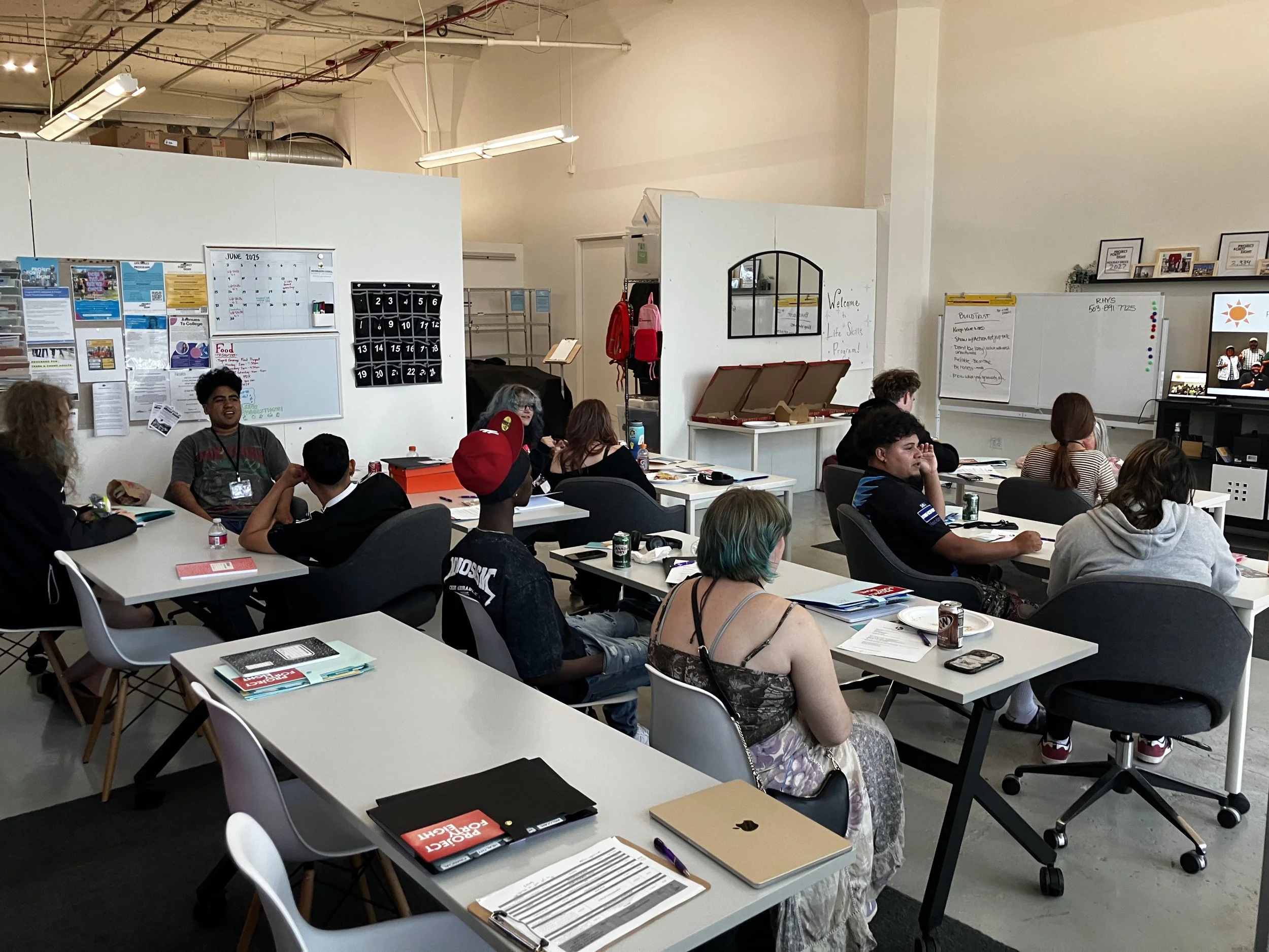A diverse group of people attending a class or meeting in a modern, open-plan space with white walls, whiteboards, and a TV screen.