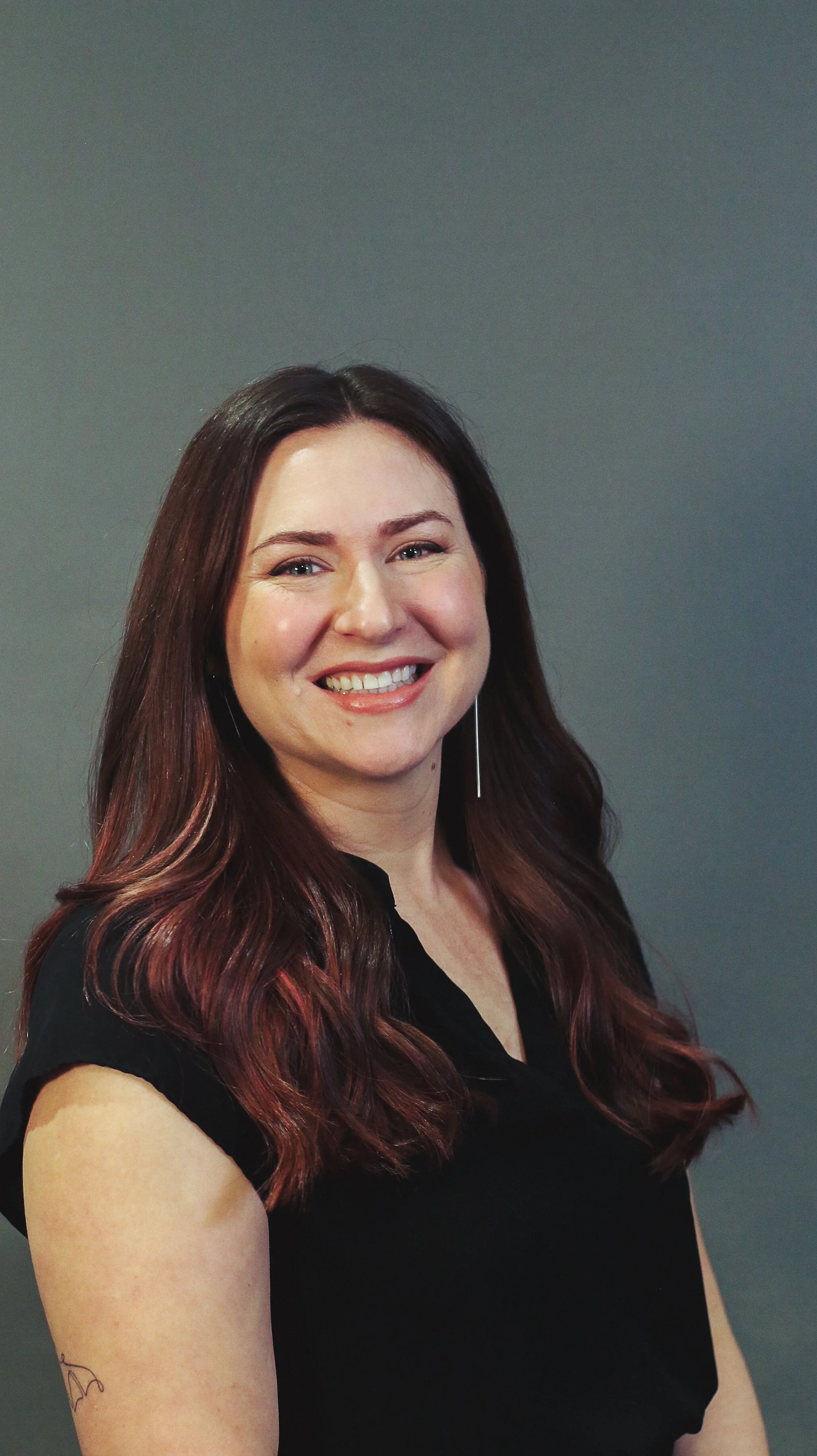 A woman with long, dark brown hair smiling, wearing a black top, and a long earring, standing against a plain gray background.