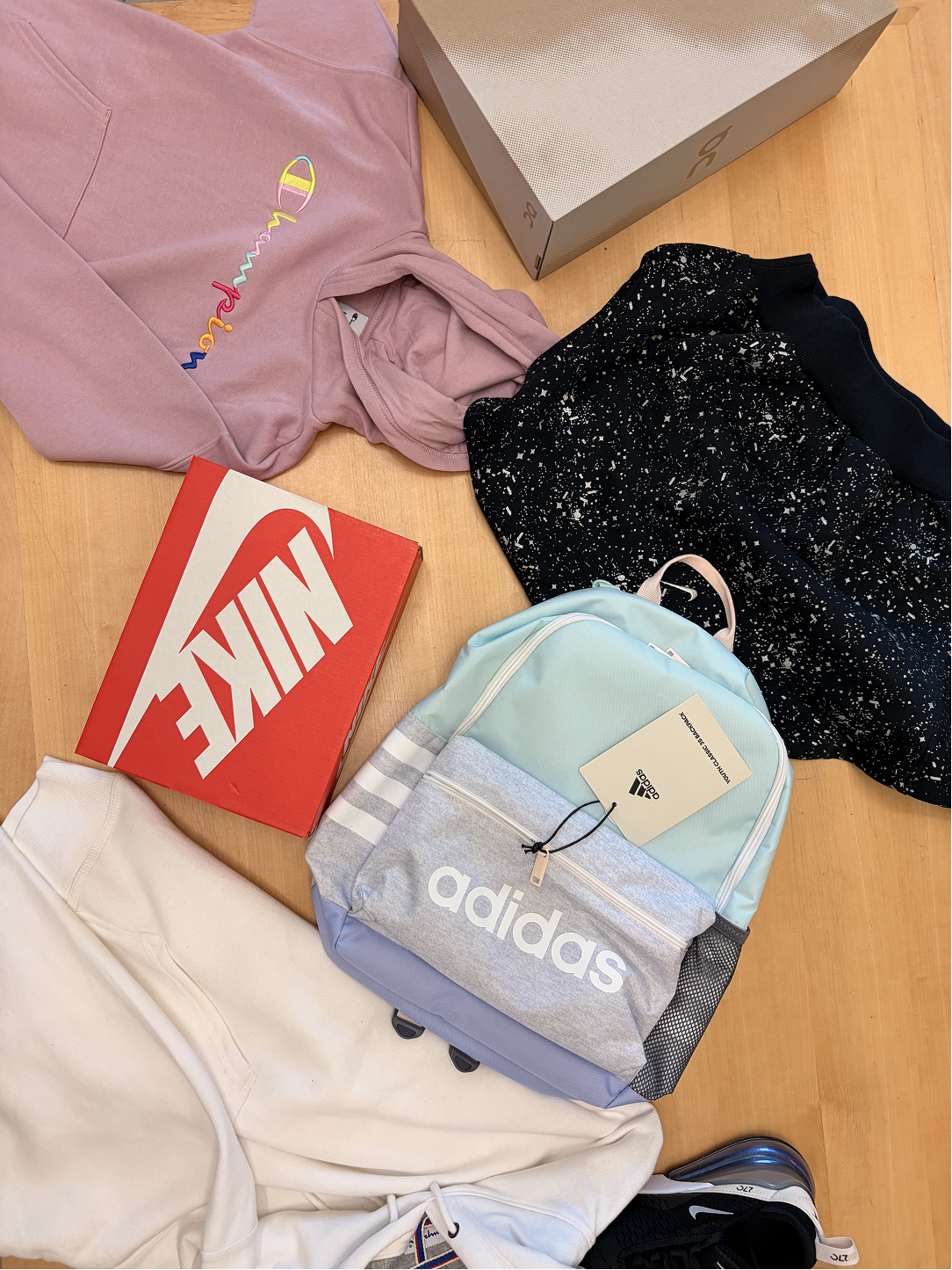 A collection of casual clothing and accessories on a wooden surface, including a pink Champion hoodie, a pair of black and white speckled shorts, a light blue Adidas backpack, a white hoodie, a red Nike shoebox, and a pair of black Nike sneakers.