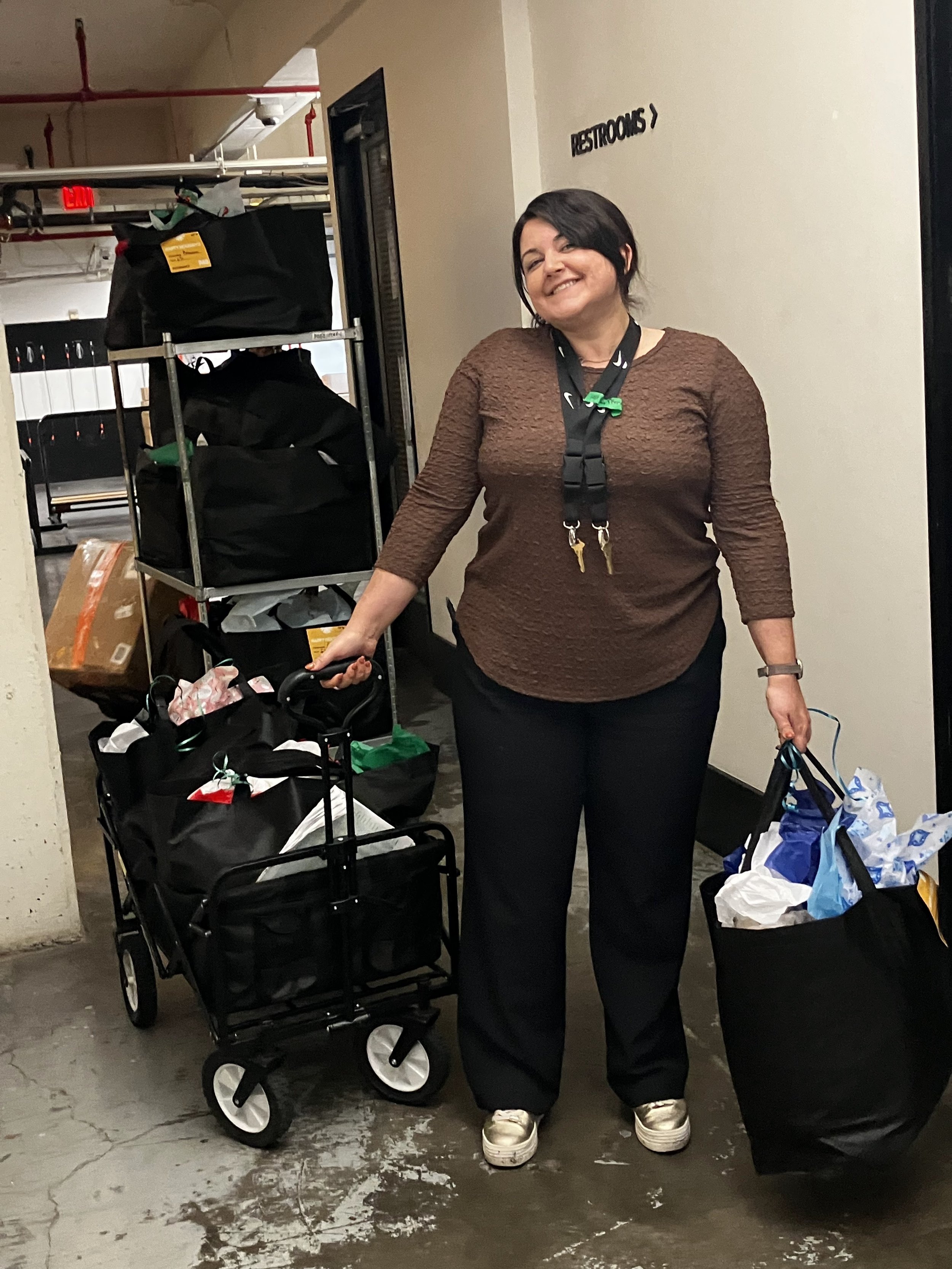 A woman standing by shopping carts in an indoor parking garage, smiling, holding a shopping cart on her left and a shopping bag in her right hand, with a sign that says 'Restrooms' on the wall behind her.