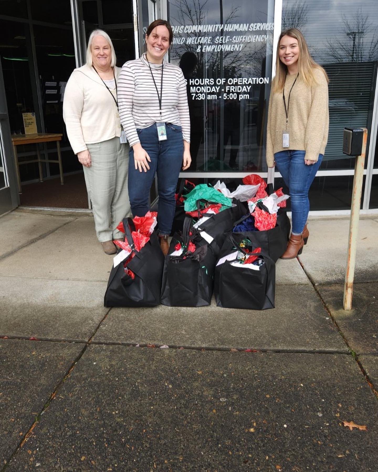 Three women standing outside a building with large black gift bags filled with holiday items on the sidewalk in front of them. They are smiling and wearing ID badges, with a glass door behind them displaying hours of operation.