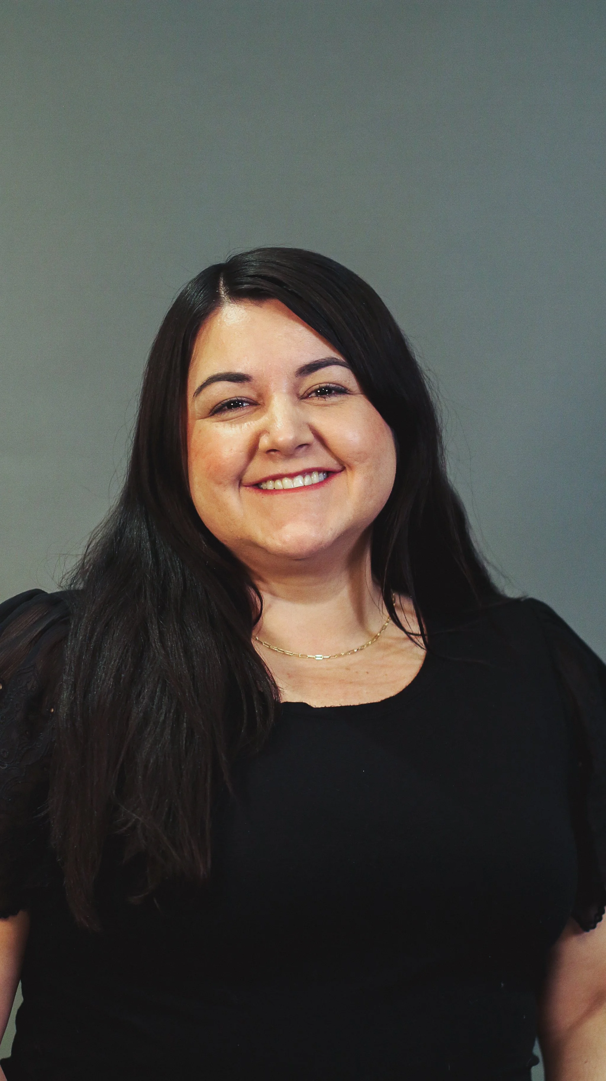 A woman with dark brown hair, wearing a black top and a thin gold necklace, smiling against a plain gray background.