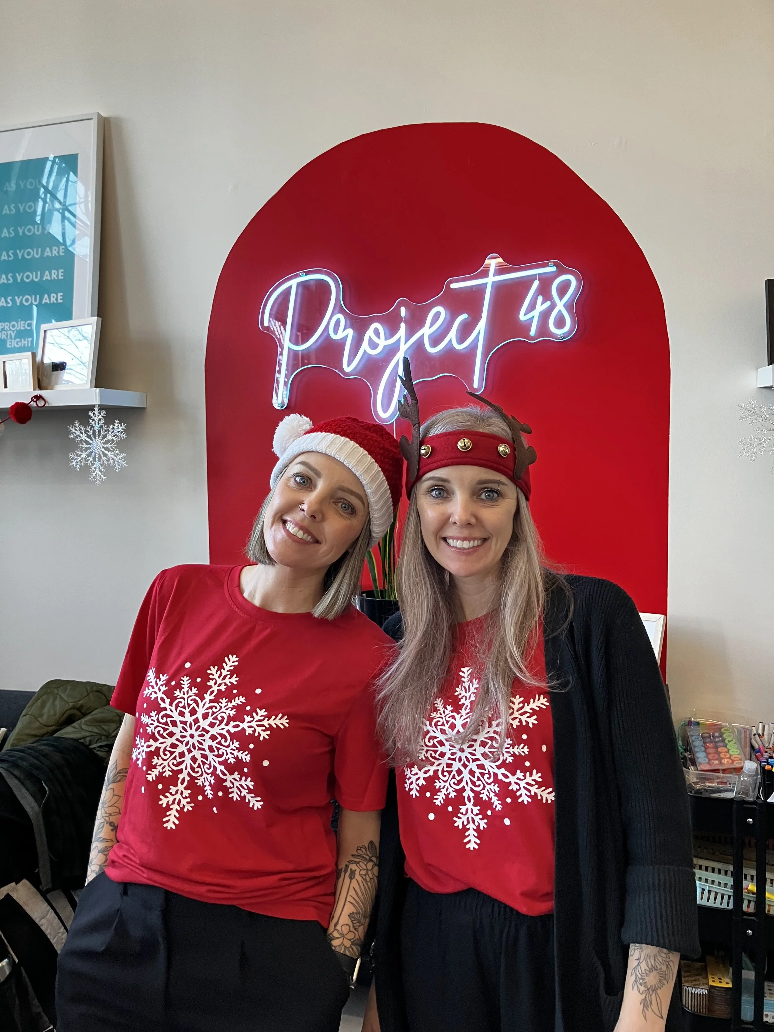Two women in Christmas-themed shirts and hats smiling in front of a neon sign that reads 'Project 48' with a red background.