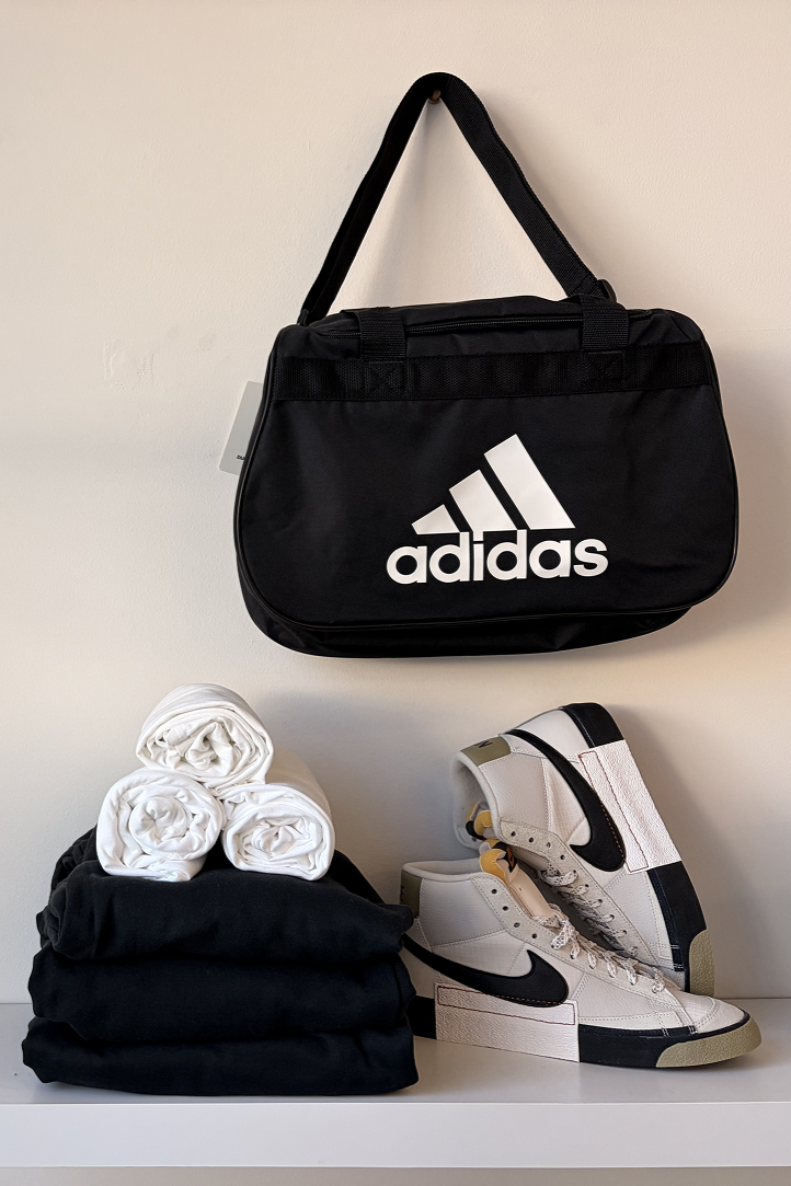 A black Adidas gym bag hanging on the wall, a stack of white rolled-up clothes or towels, black pants, and white Nike high-top sneakers with black swoosh and beige accents on a white surface.