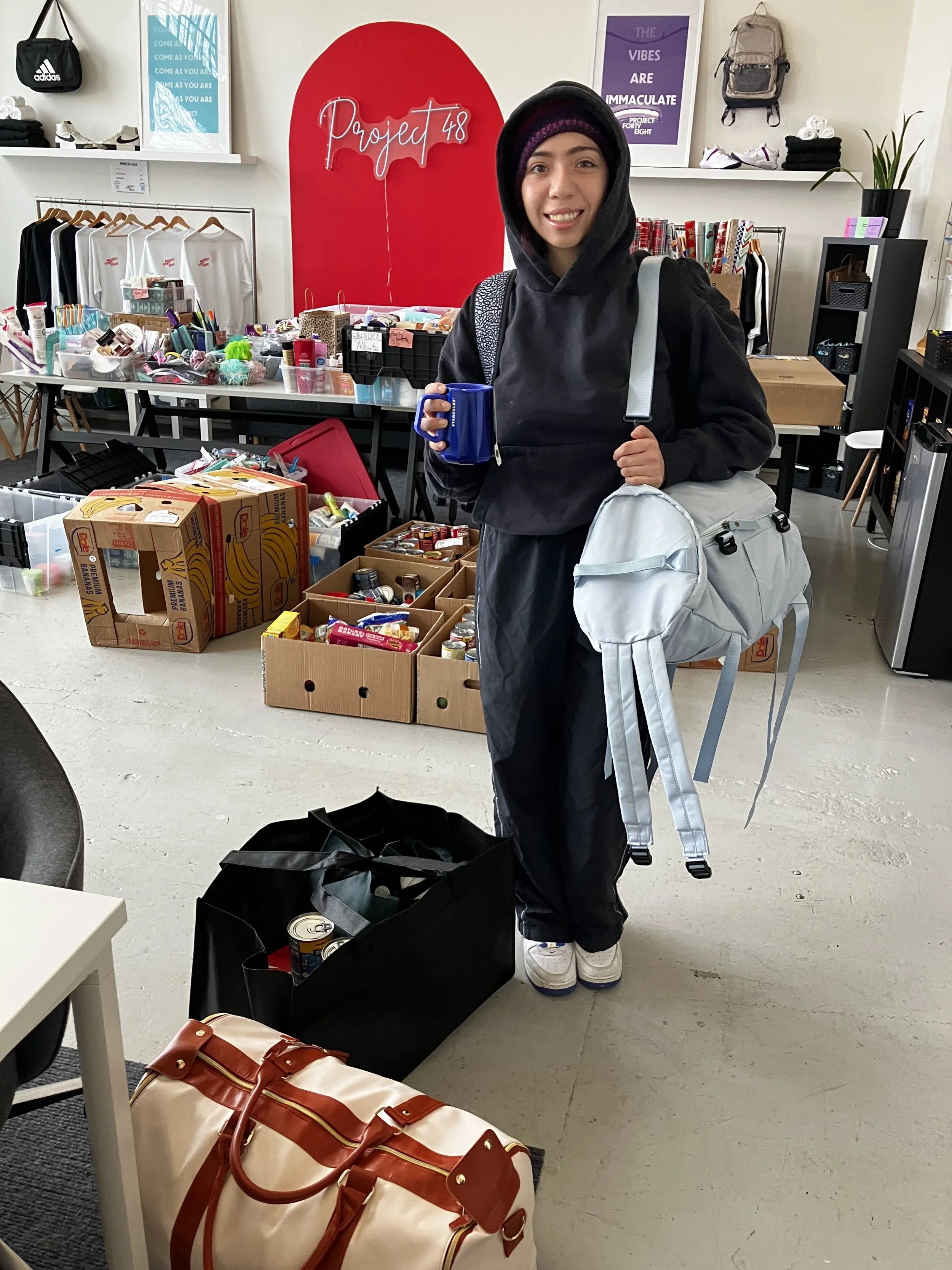 A young woman with a backpack and a blue coffee mug standing in a store or donation center, surrounded by boxes of canned goods and groceries, with shelves of clothing and motivational posters in the background.