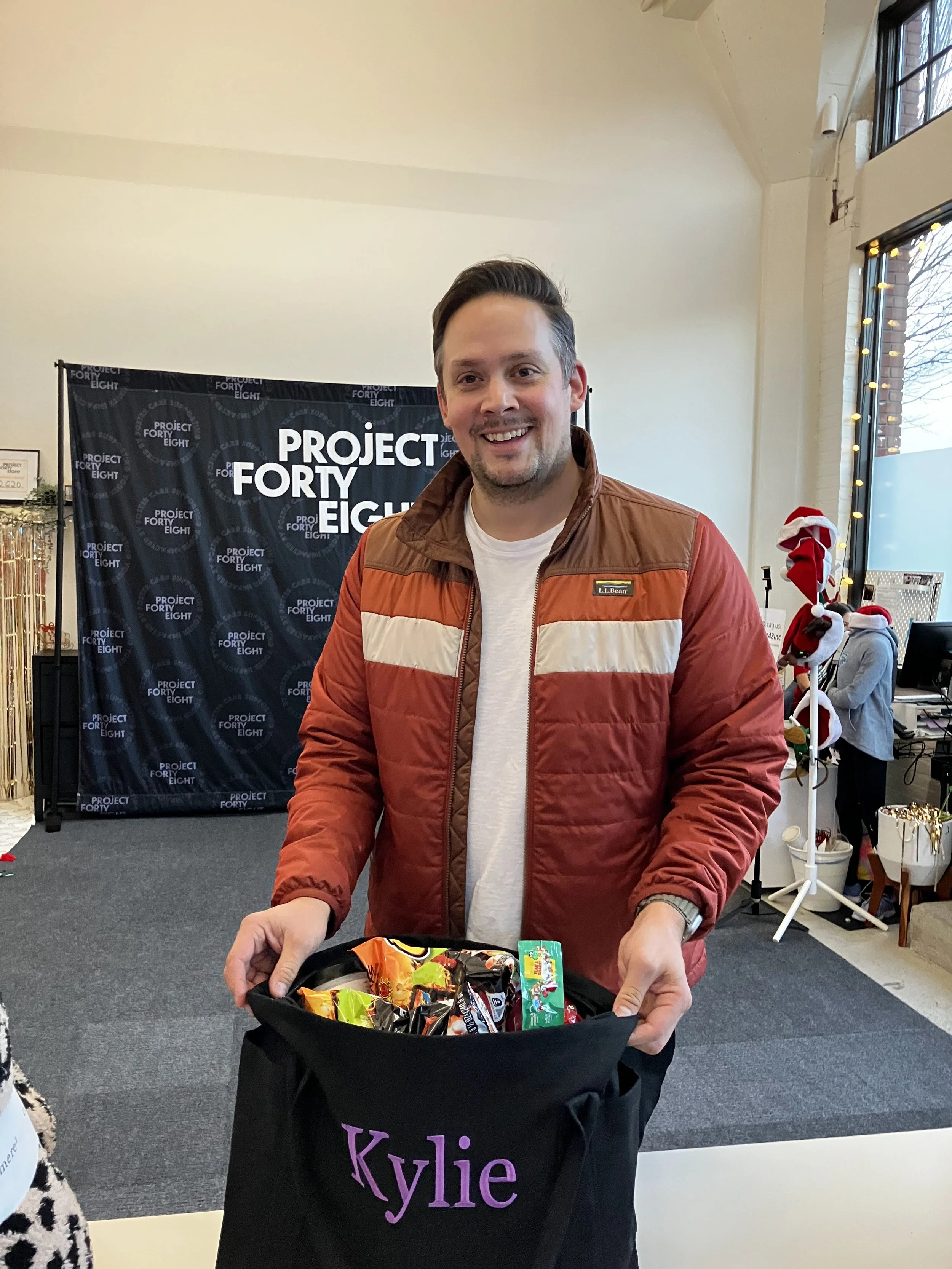 A smiling man holding a bag of snacks labeled 'Kylie' in a room decorated for Christmas, with a 'Project Forty Eight' backdrop and holiday decorations.