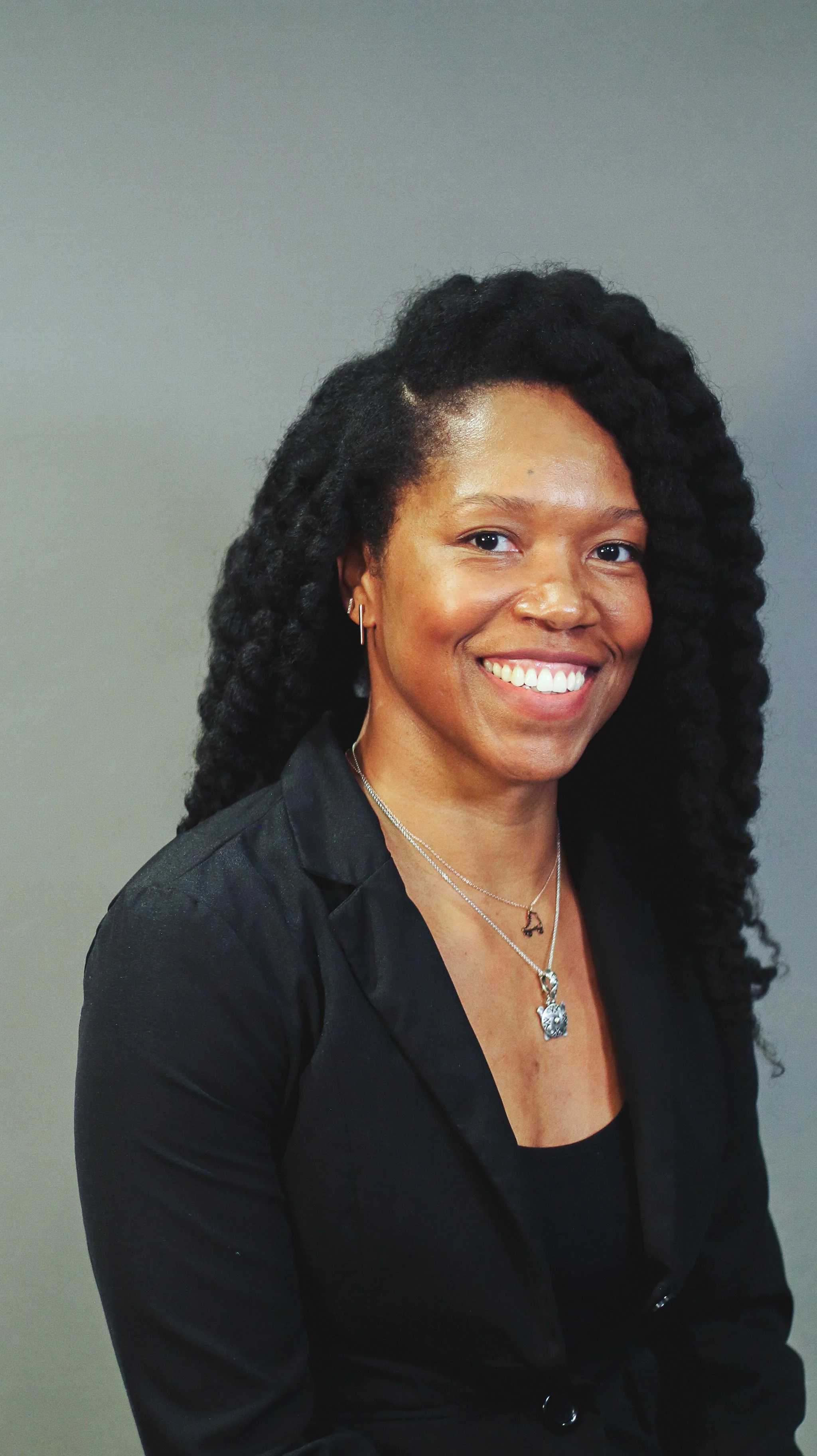 A smiling woman with dark, curly hair wearing a black blazer and layered necklaces, standing against a plain gray background.