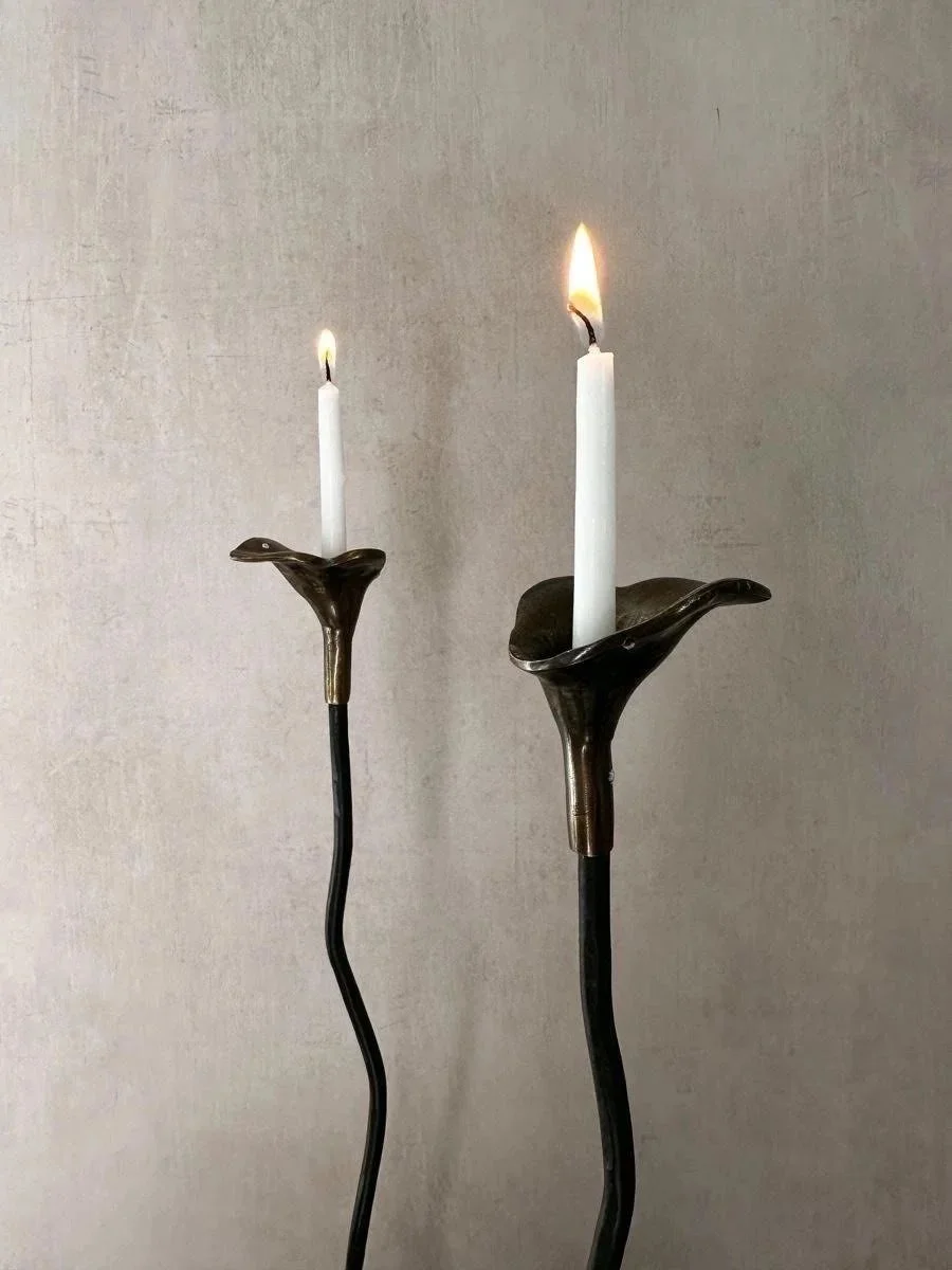 Two modern candle holders with white candles, one lit and the other unlit, against a plain wall.