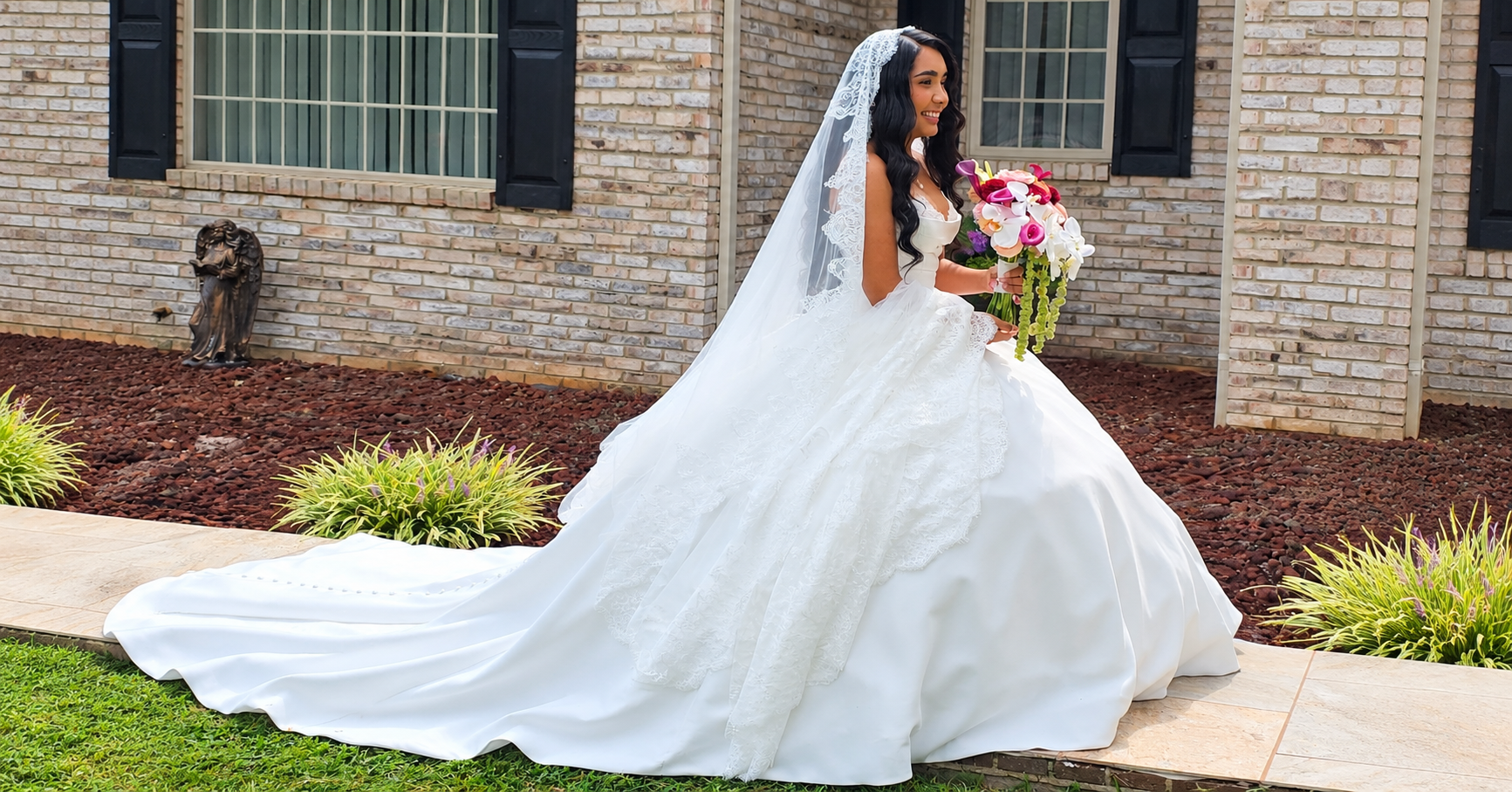 Bride wearing a lace veil with long black curls and a white wedding gown, holding a colorful bouquet.