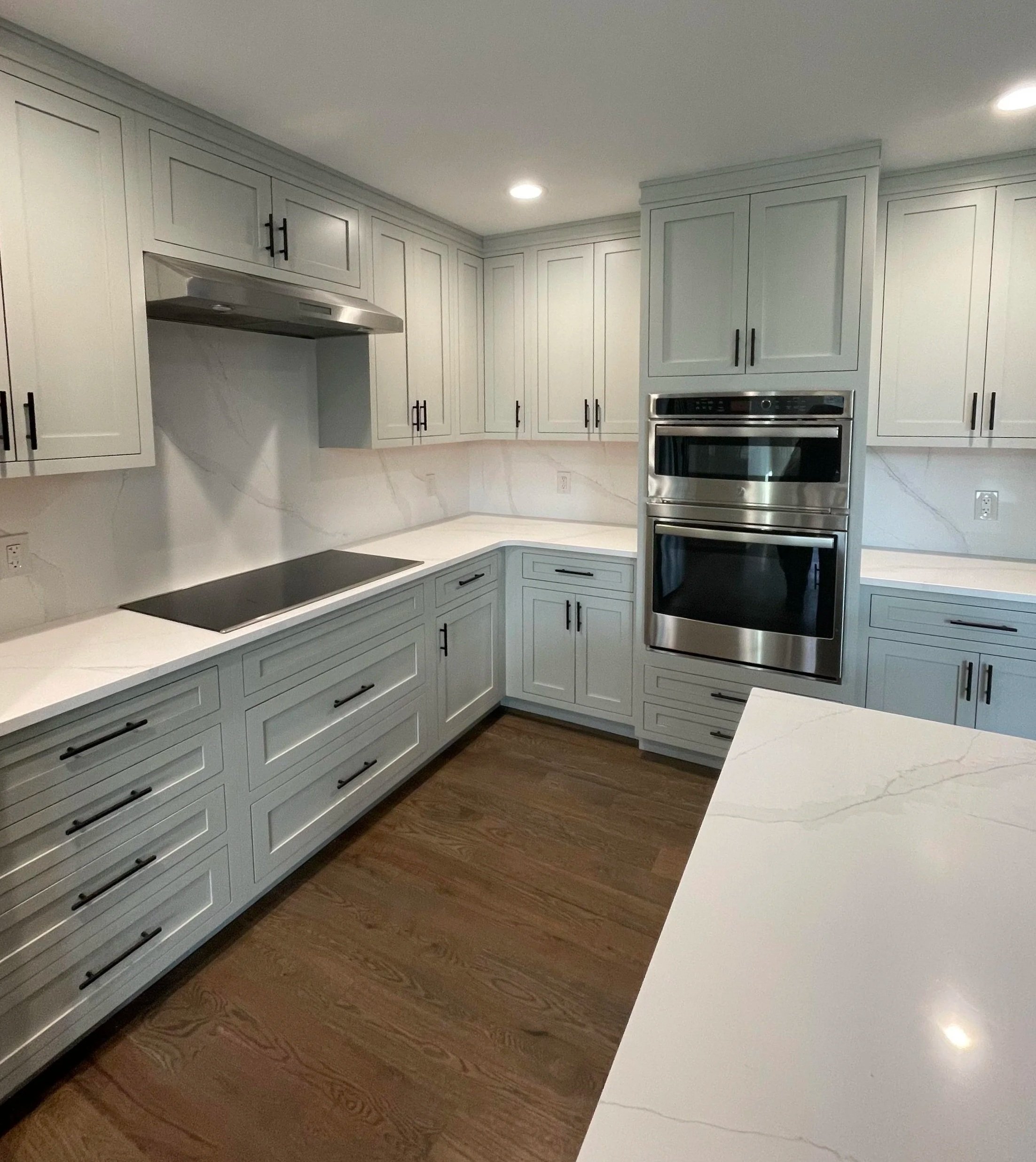 A modern kitchen with white cabinets, black hardware, a built-in stainless steel oven, a black stovetop, white marble countertops, and hardwood floors.
