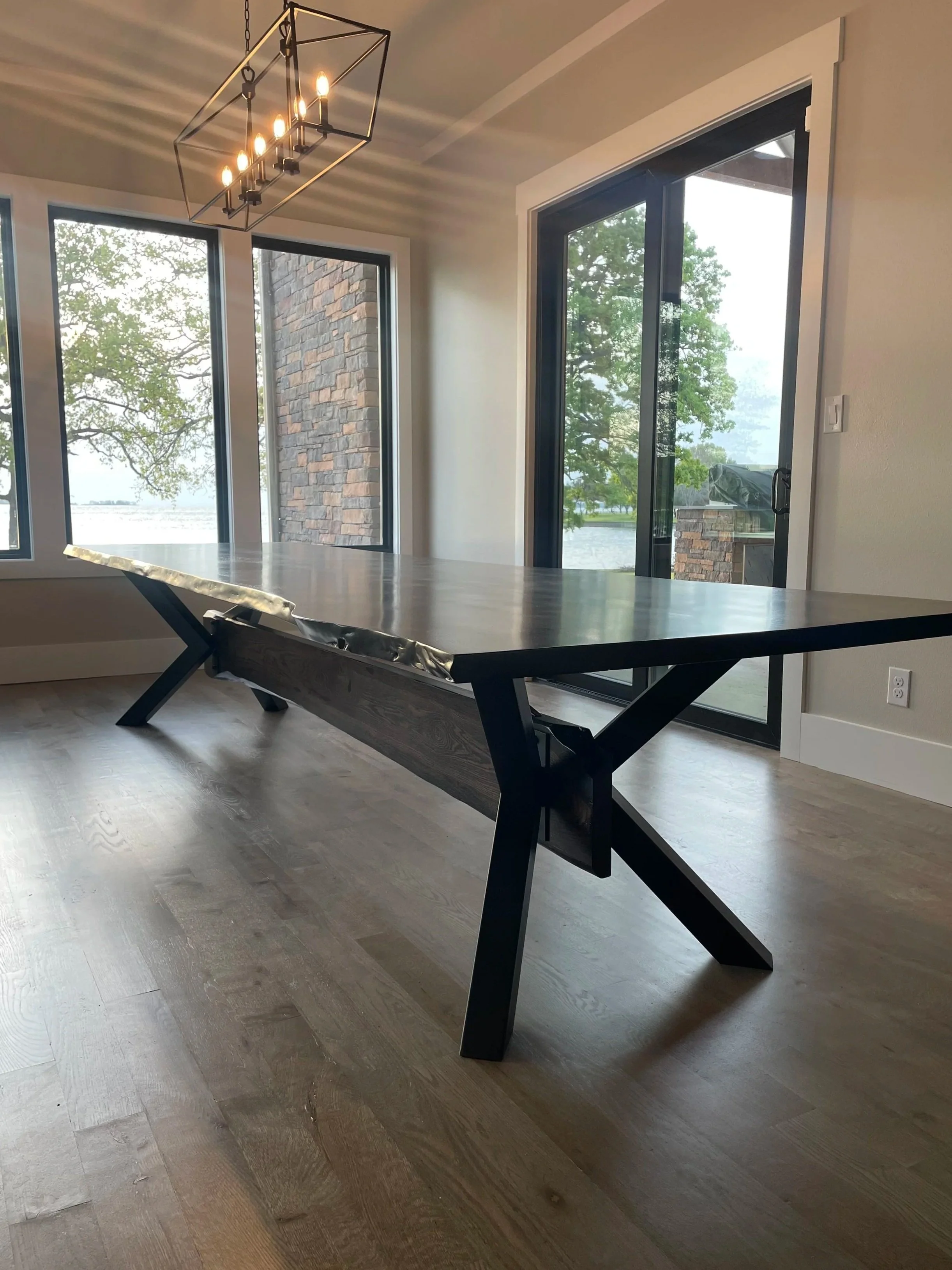 A modern dining room with a black dining table, large windows showing trees and a body of water, and a geometric black chandelier with six lights hanging from the ceiling.