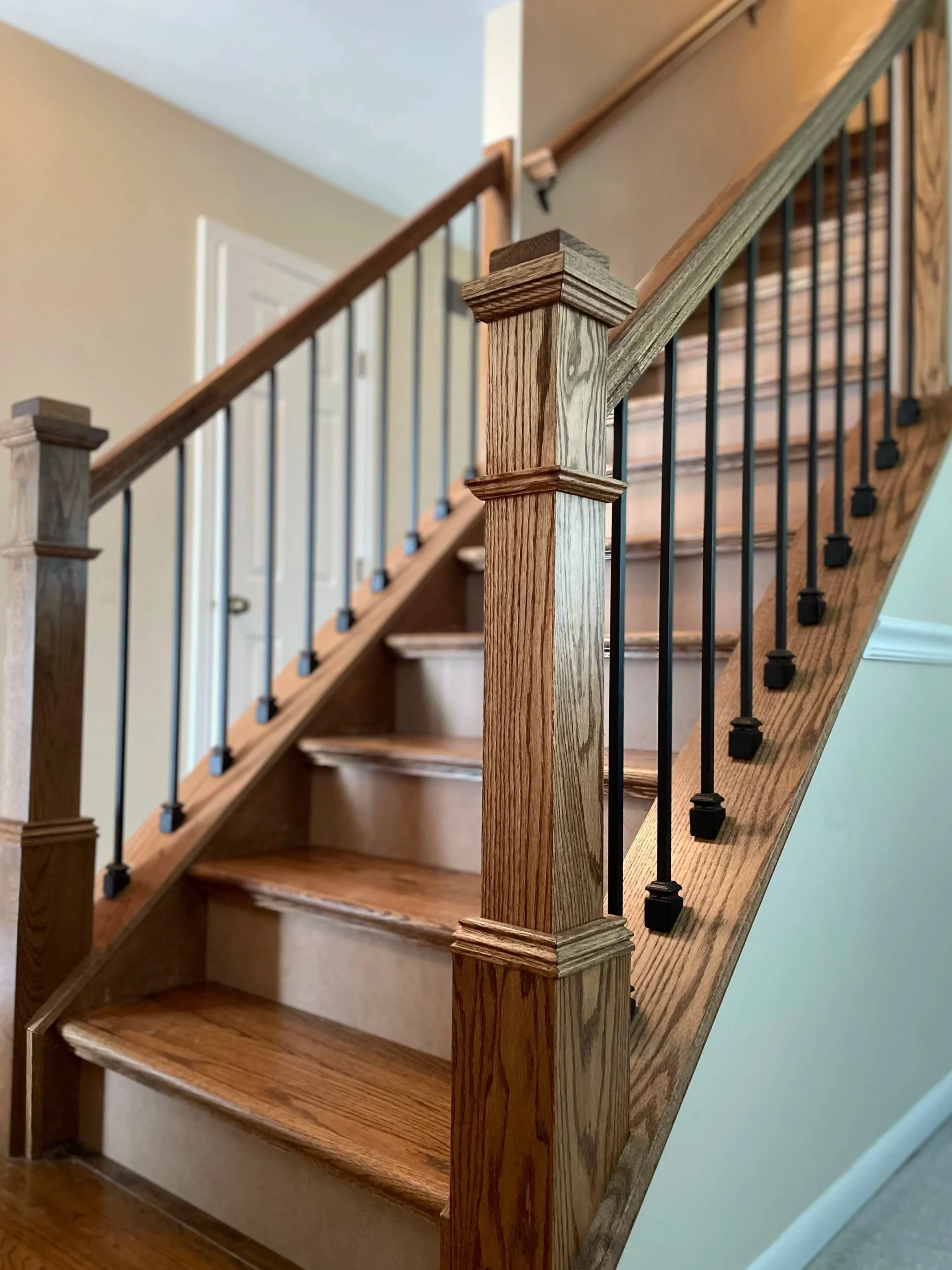 Wood staircase with black metal balusters and wooden handrails and newel posts, carpeted stairs in the background, inside a house.