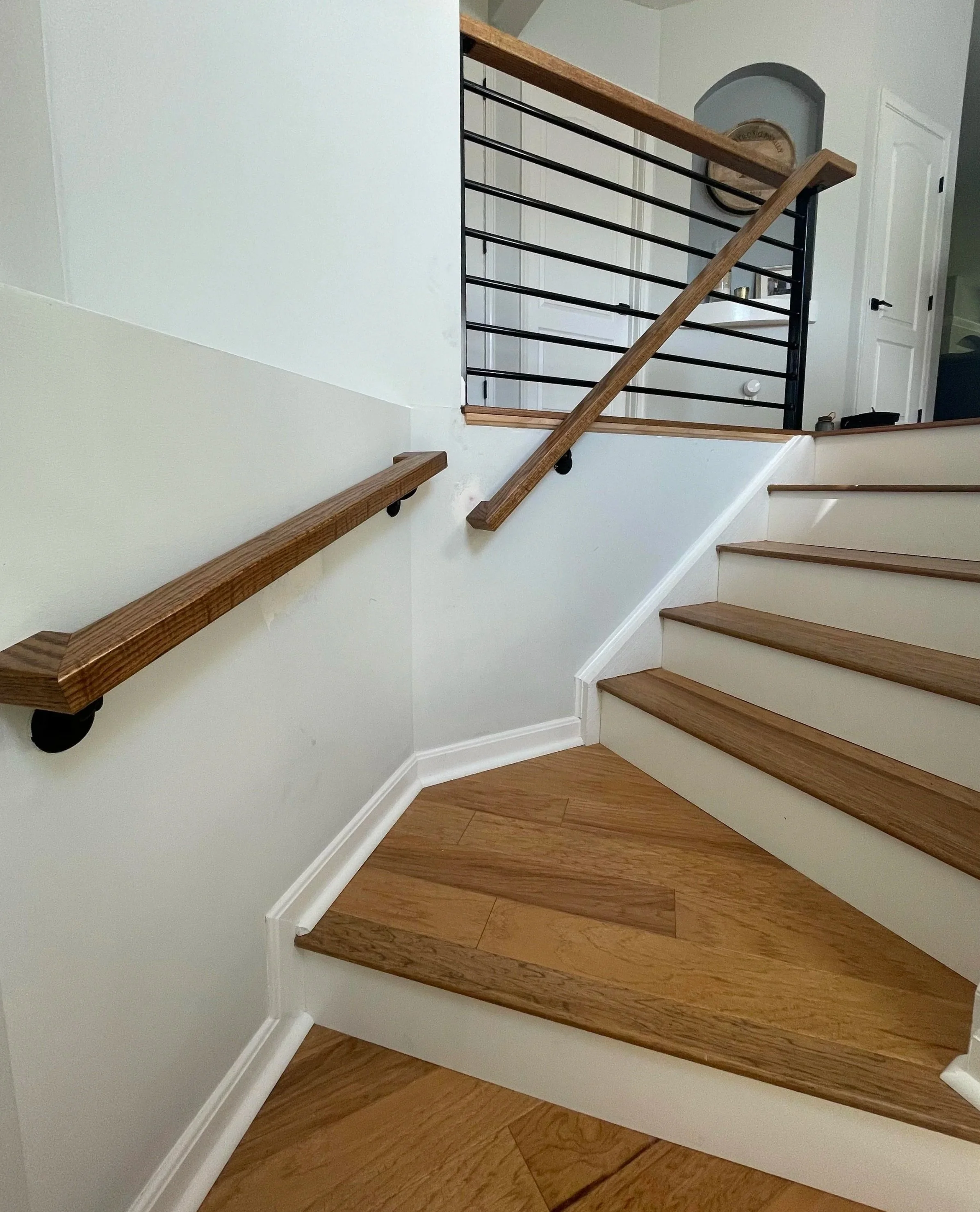 Indoor staircase with wooden handrails, white risers, and wood-look steps, along with a black metal railing on the upper level. White walls and a door are visible in the background.