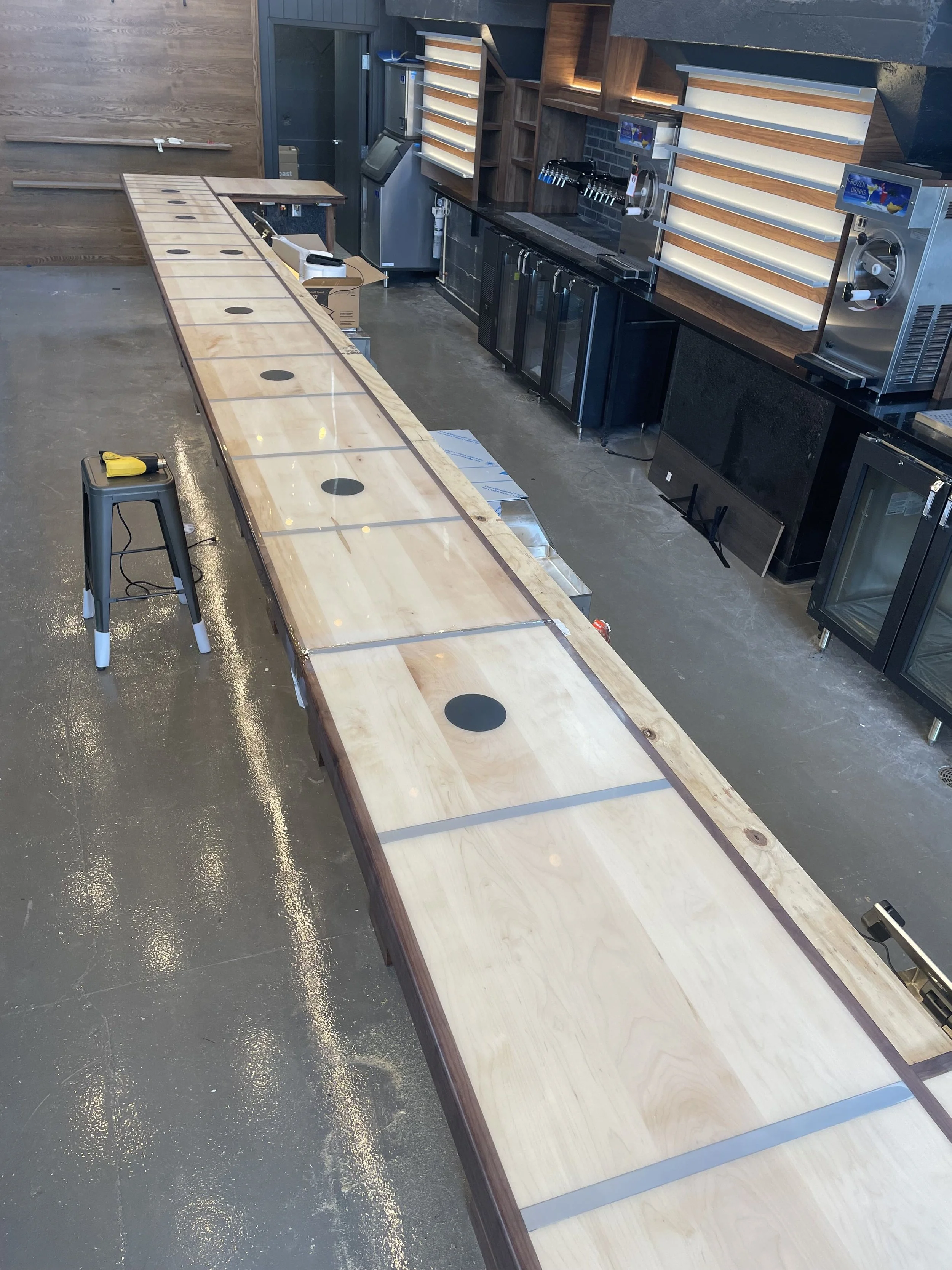 A long wooden table in a commercial space with a stool at one end, and bar equipment along the wall including shelves, refrigerators, and dispensers.