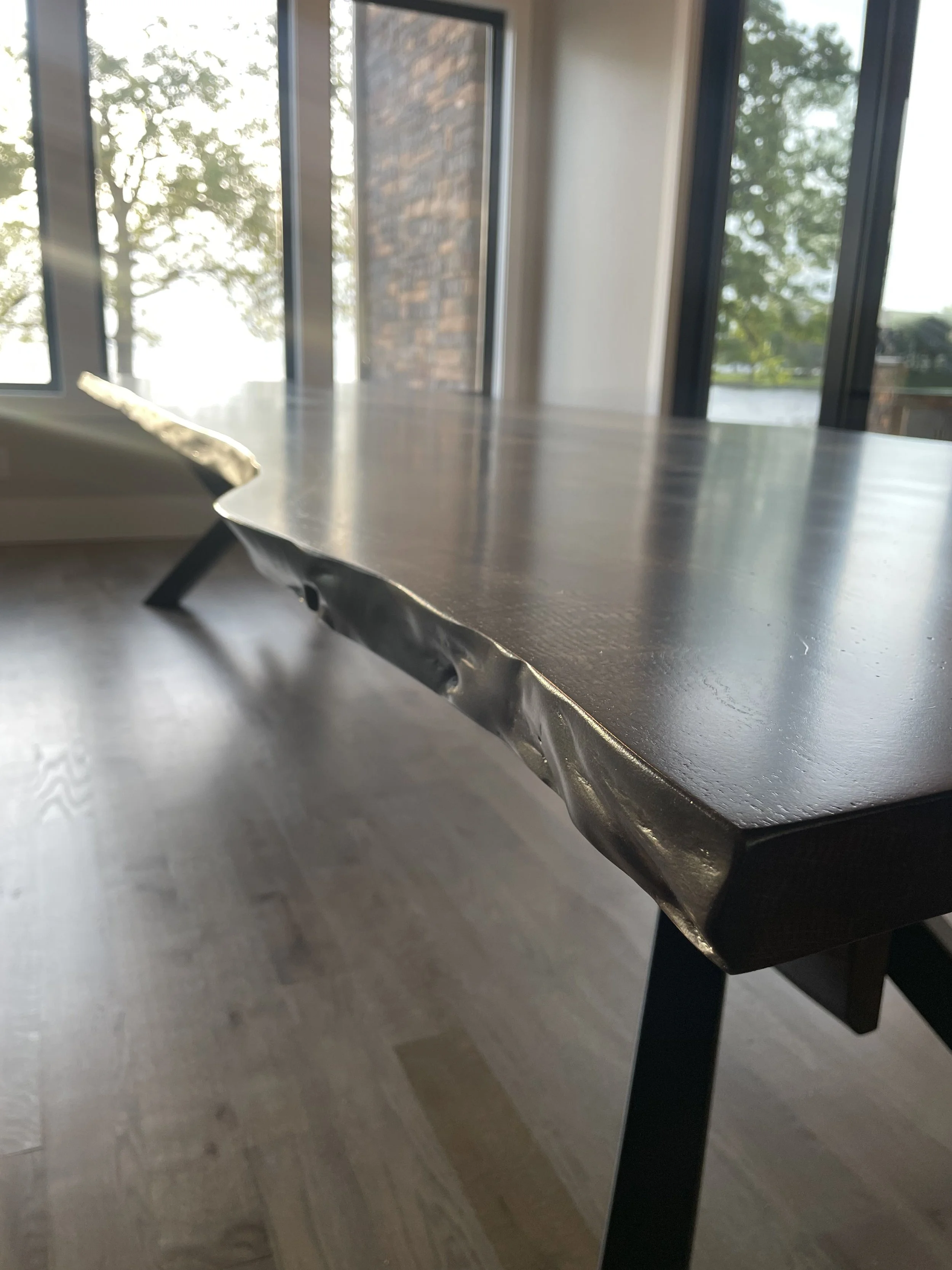 Close-up of a dark wooden table with a metal edge and a black leg, beside large glass windows showing trees and brick walls outside.