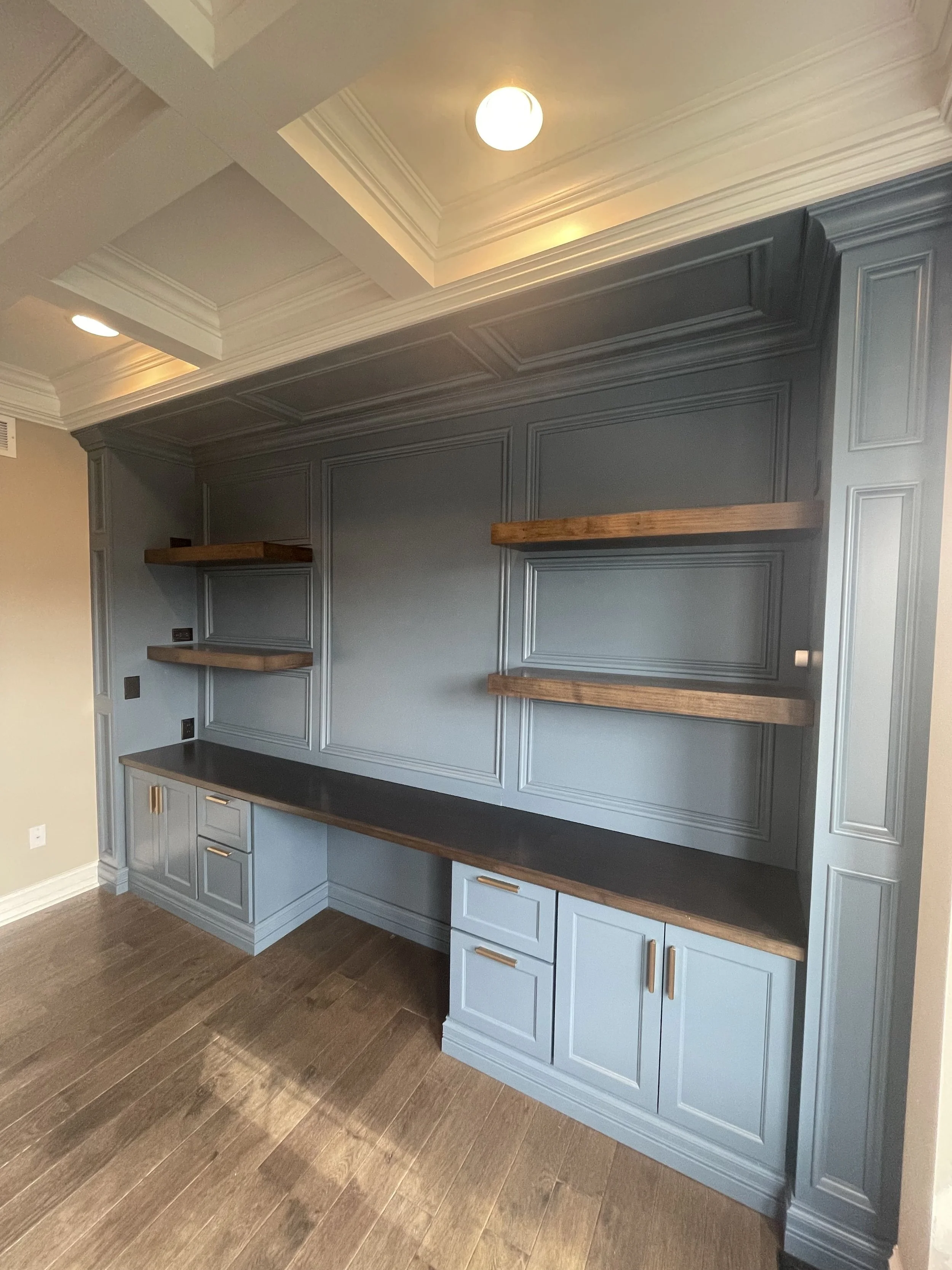 Interior view of a built-in cabinet with dark gray paint and wooden open shelves, set against a wall with decorative molding, above a wooden countertop, with a ceiling light and recessed lighting, and hardwood flooring.