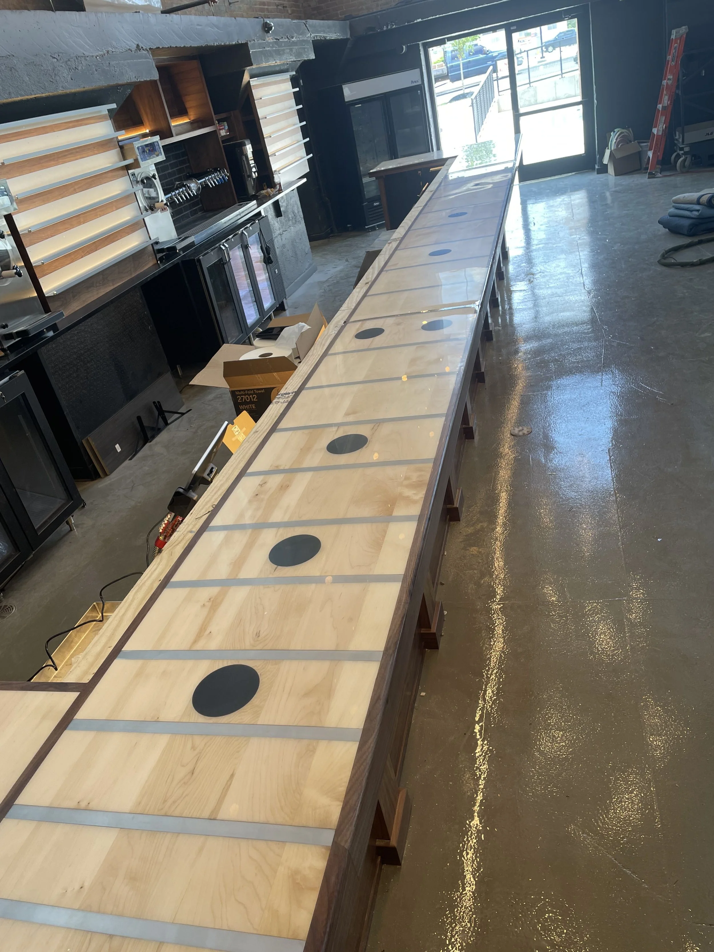 Interior of a commercial space under construction with a long wooden countertop, black appliance cabinets, and a large window or glass door at the end revealing a parking lot outside.