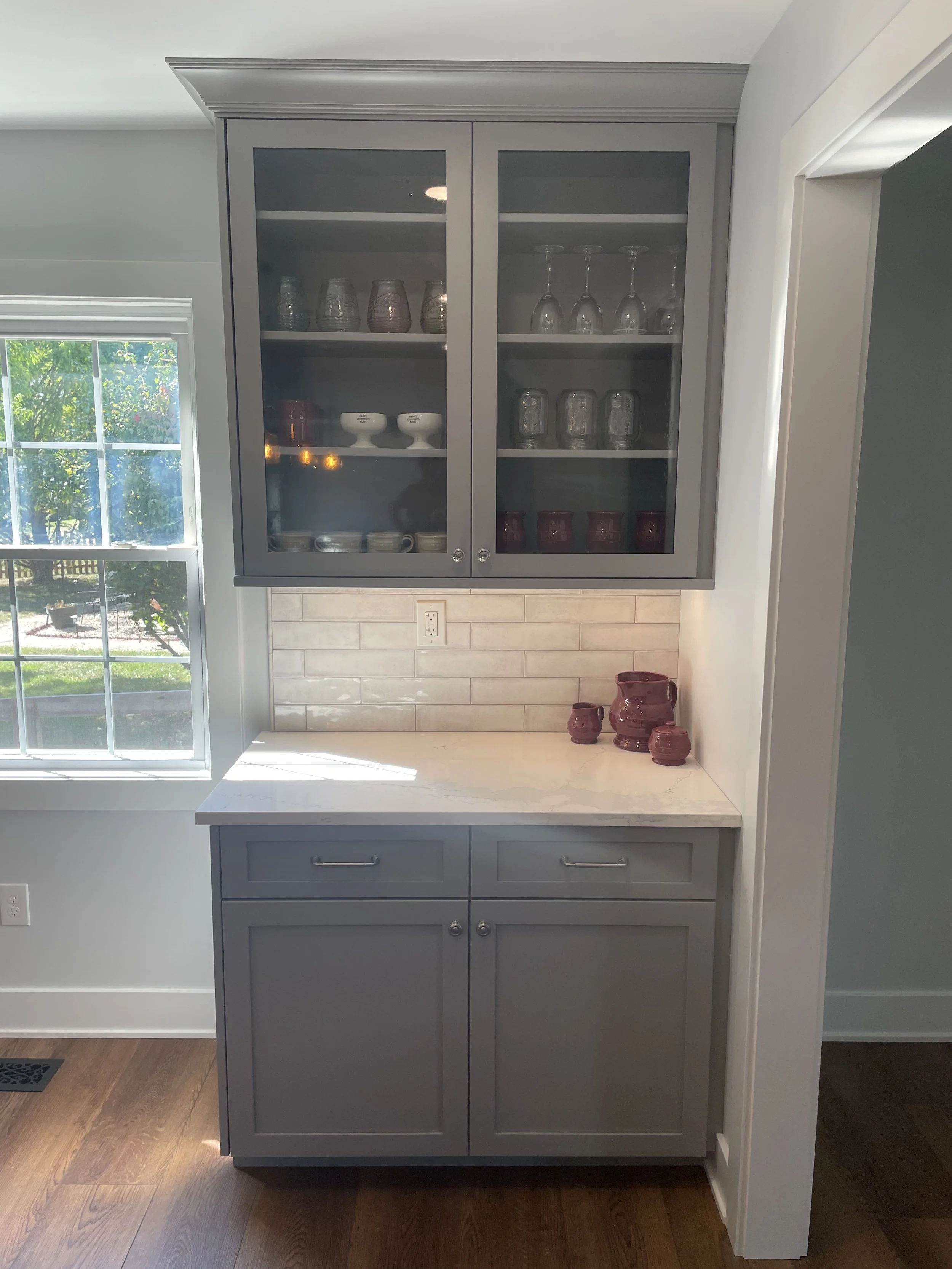 A kitchen cabinet with glass doors displaying glassware and dishes, placed above a countertop with decorative red vases, next to a window with sunlight and a view of a garden outside.