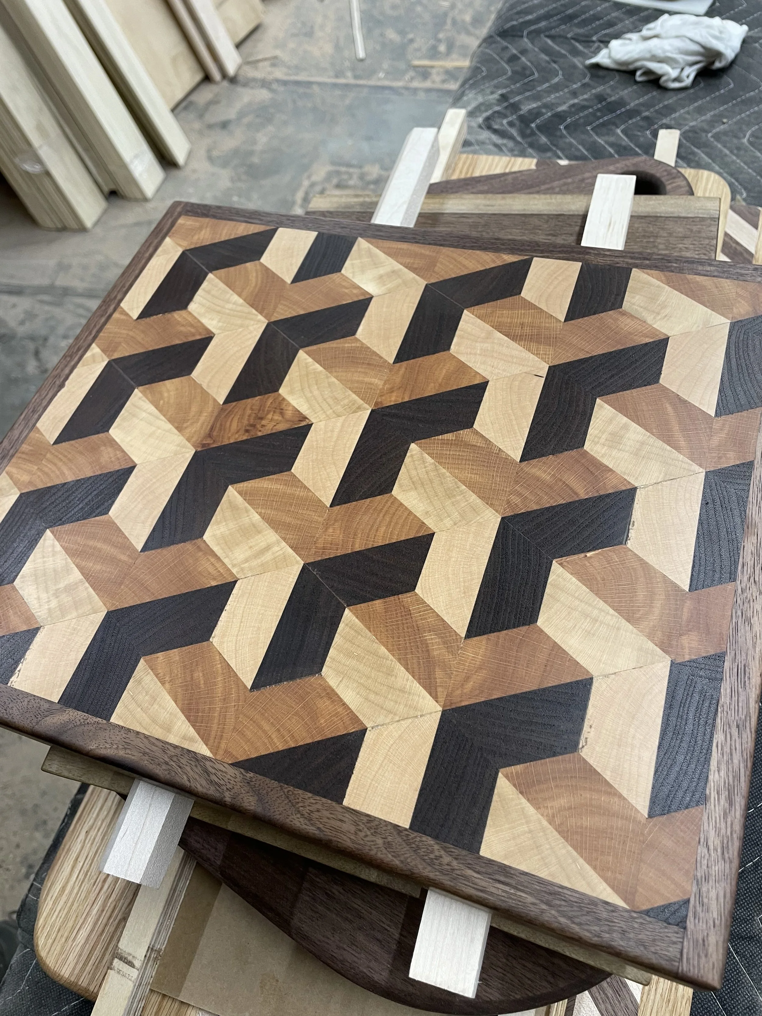 A wooden table or panel featuring a geometric pattern design made of different types and shades of wood, with some pieces extending beyond the edge. The background includes a workshop setting with wood pieces, a crumpled cloth, and a black cloth with