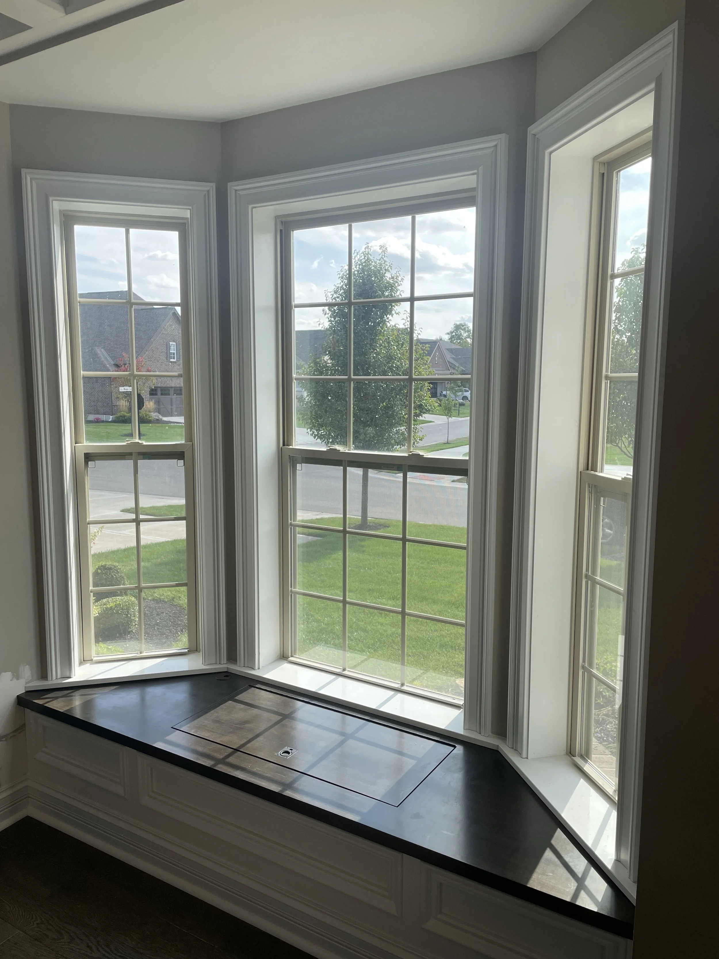 Interior view of three large windows with white trim, overlooking a residential neighborhood with houses, trees, and a street, with a black countertop beneath the windows.