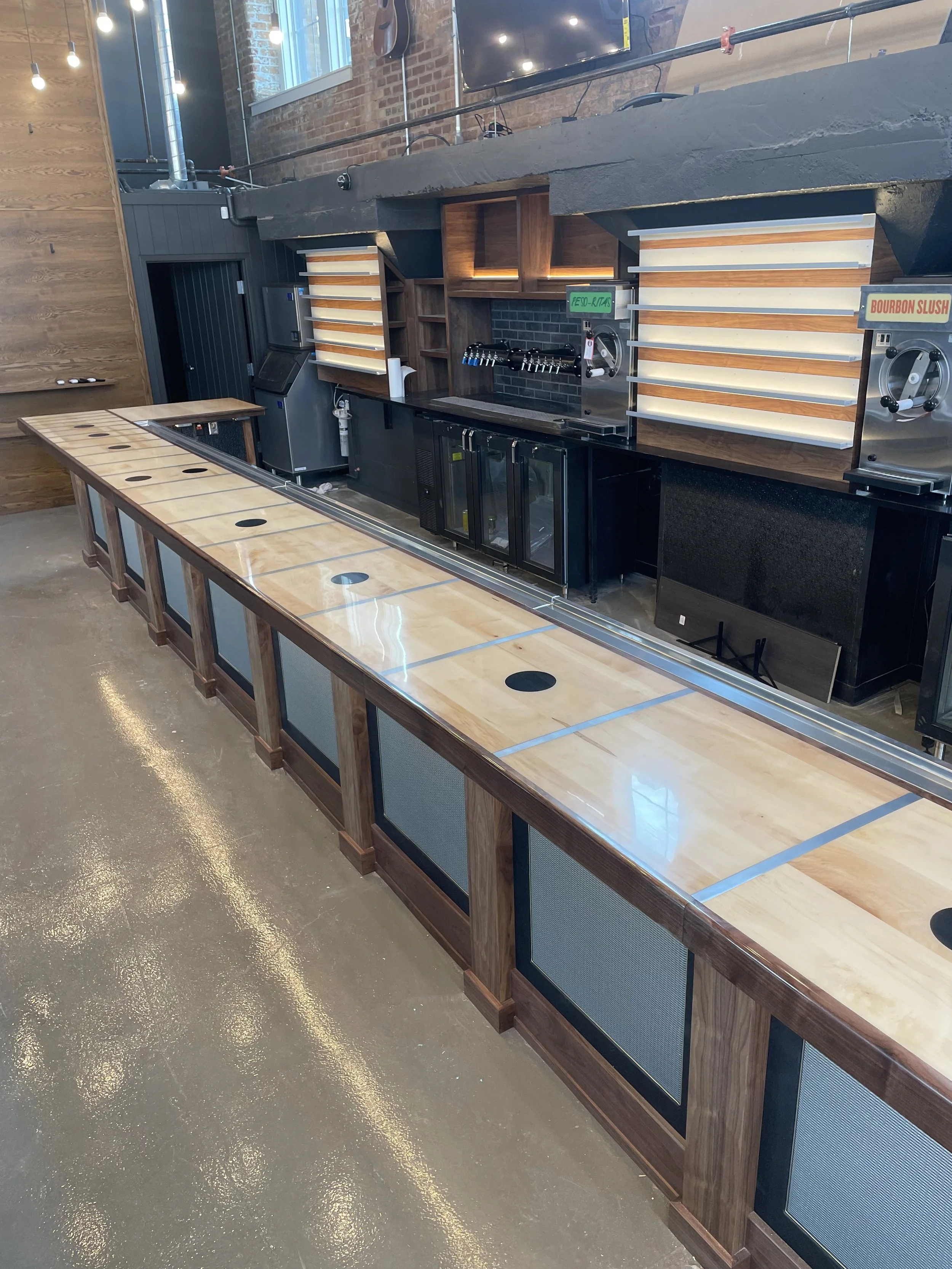 A long wooden and black bar counter with no stools, set in a modern bar or restaurant. The bar has insets with black panels, and the countertop has hole covers for drink dispensers. Behind the counter are shelves with liquor bottles, a beer tap, and 