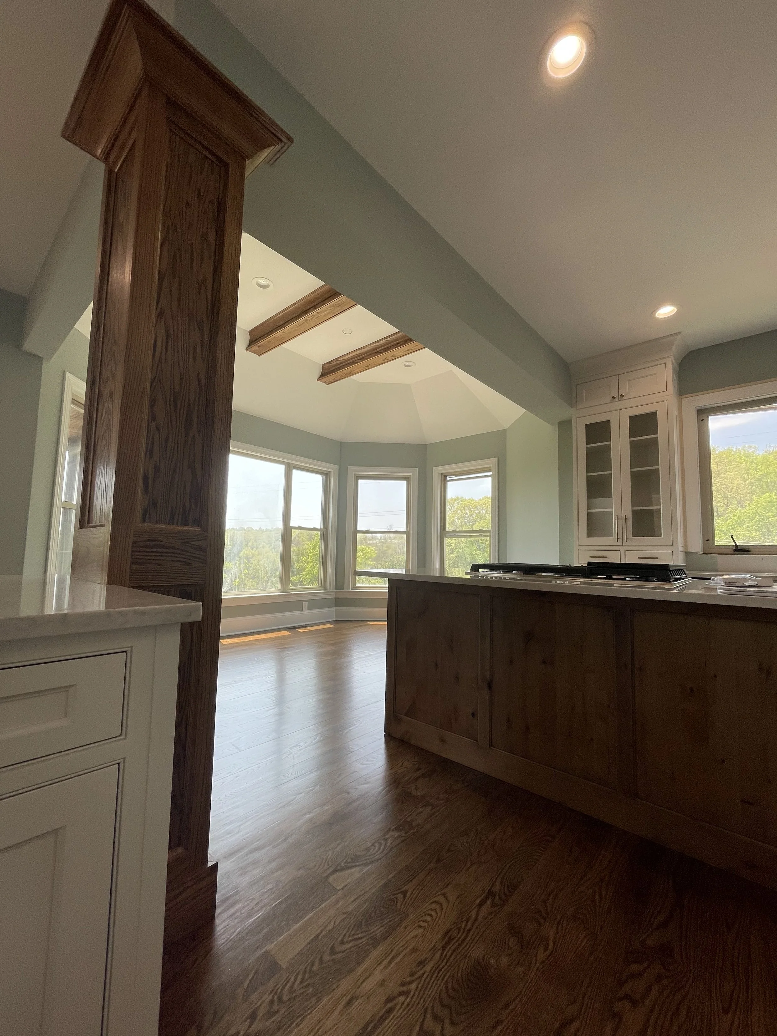 Interior view of a kitchen with large bay windows, wooden floors, white cabinetry, and a kitchen island with a cooktop. The ceiling has recessed lighting and decorative wooden beams.