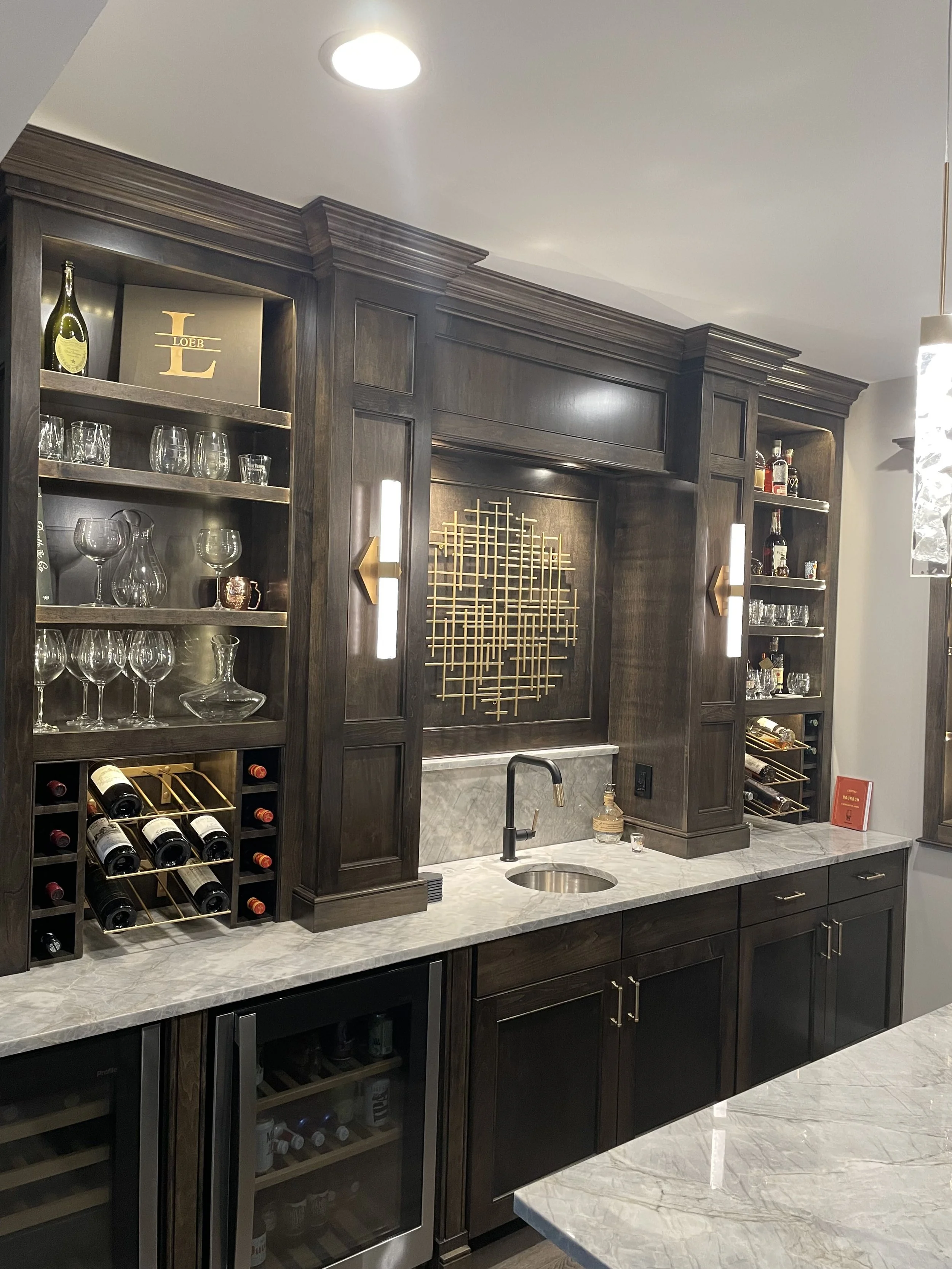 A luxurious bar area with dark wood cabinetry, open shelving with glassware and bottles, a marble countertop with a sink, and under-counter wine beverage refrigerators.