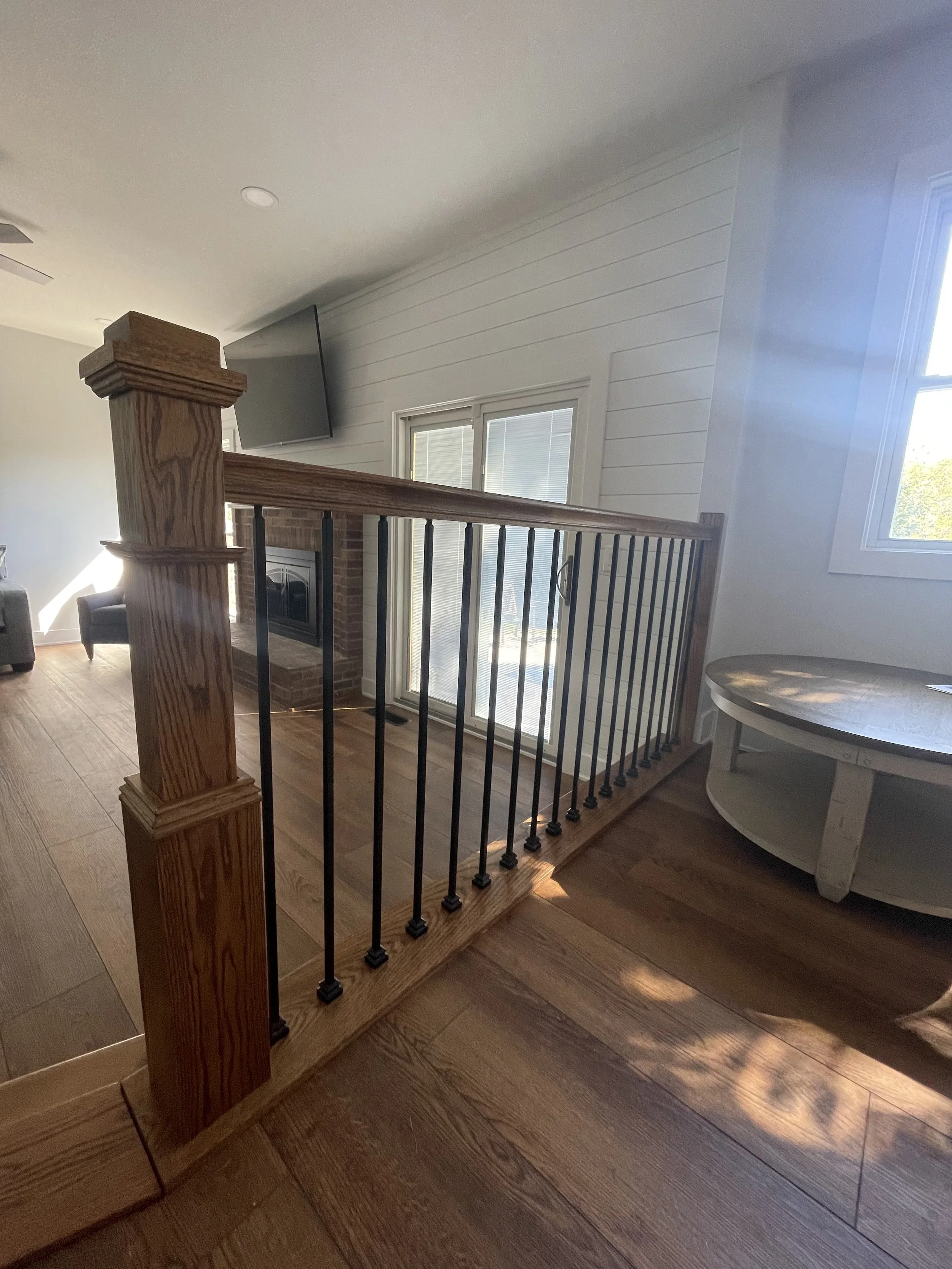 Interior of a home with a staircase featuring a wooden handrail and black metal balusters, a brick fireplace, a sitting chair, and large windows letting in natural light.